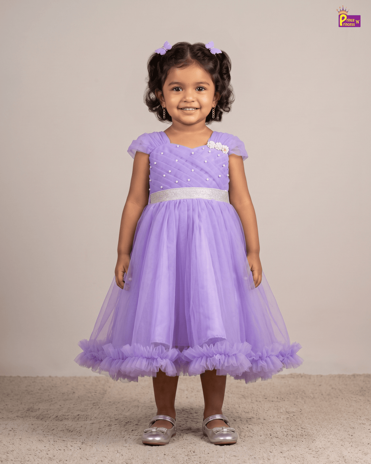 A young girl with curly dark hair wearing a lavender purple tulle party dress with a silver sash and silver shoes, standing against a plain beige studio background.