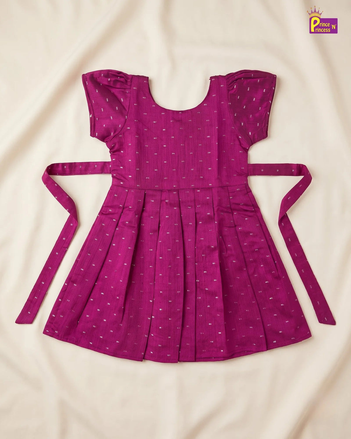 A magenta purple cotton dress with a small white pattern, featuring flutter sleeves, a pleated skirt, and matching tie straps, laid flat on a cream-colored fabric background.