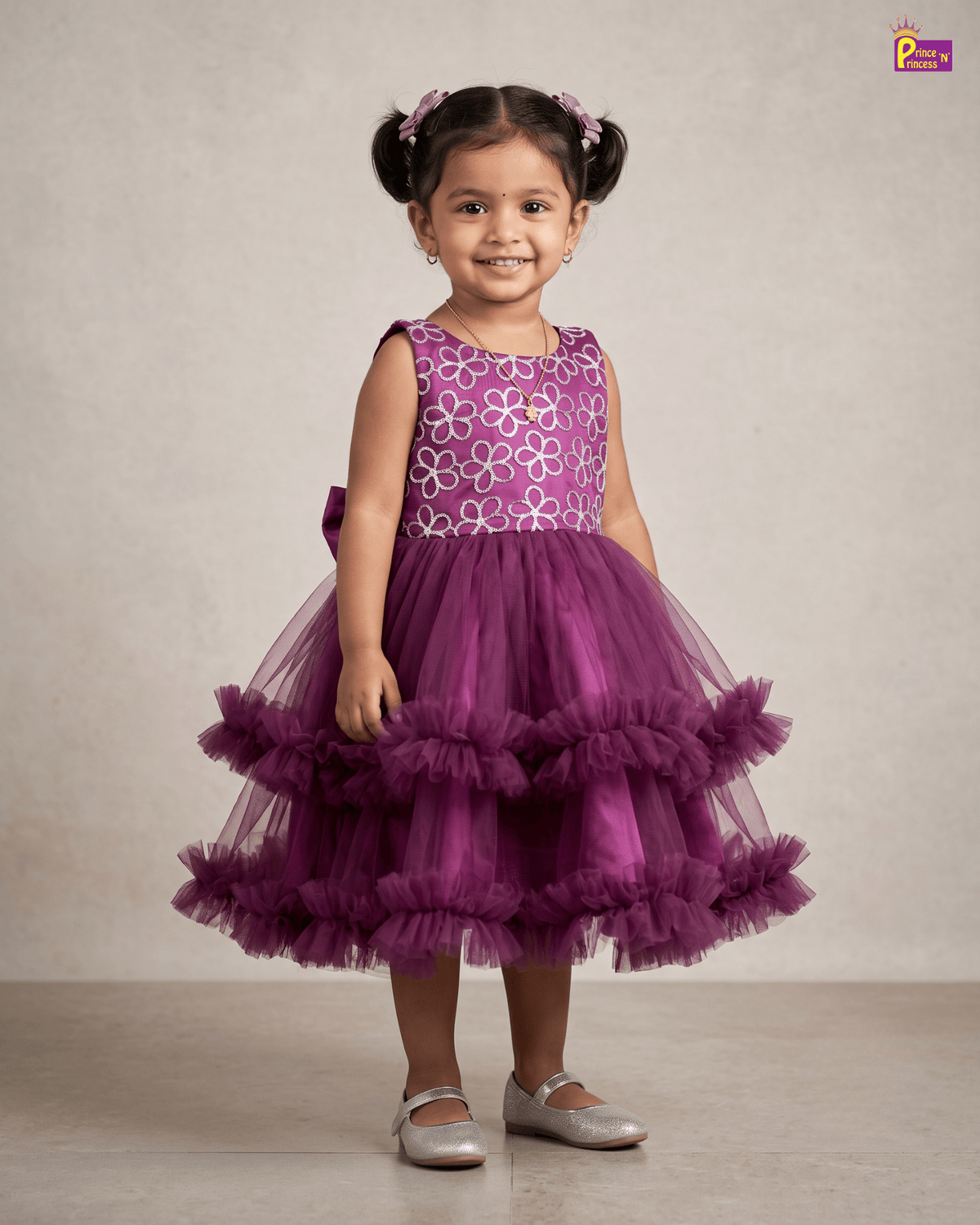 A young girl with her hair in two buns stands wearing a sleeveless purple dress with a white floral pattern on the bodice and a tiered tulle skirt. She is wearing silver ballet flats against a neutral beige studio background.