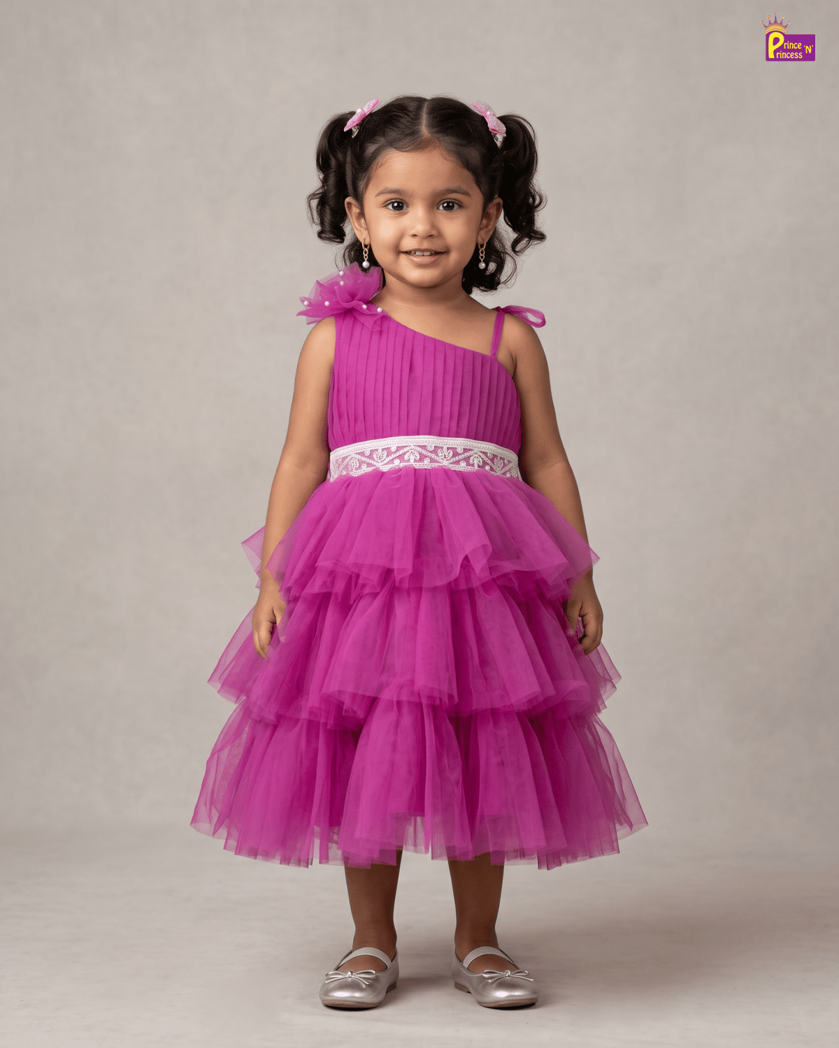 A young girl with dark curly hair in pigtails wearing a bright magenta pink one-shoulder tulle dress with a white lace waistband, standing against a neutral beige studio backdrop.