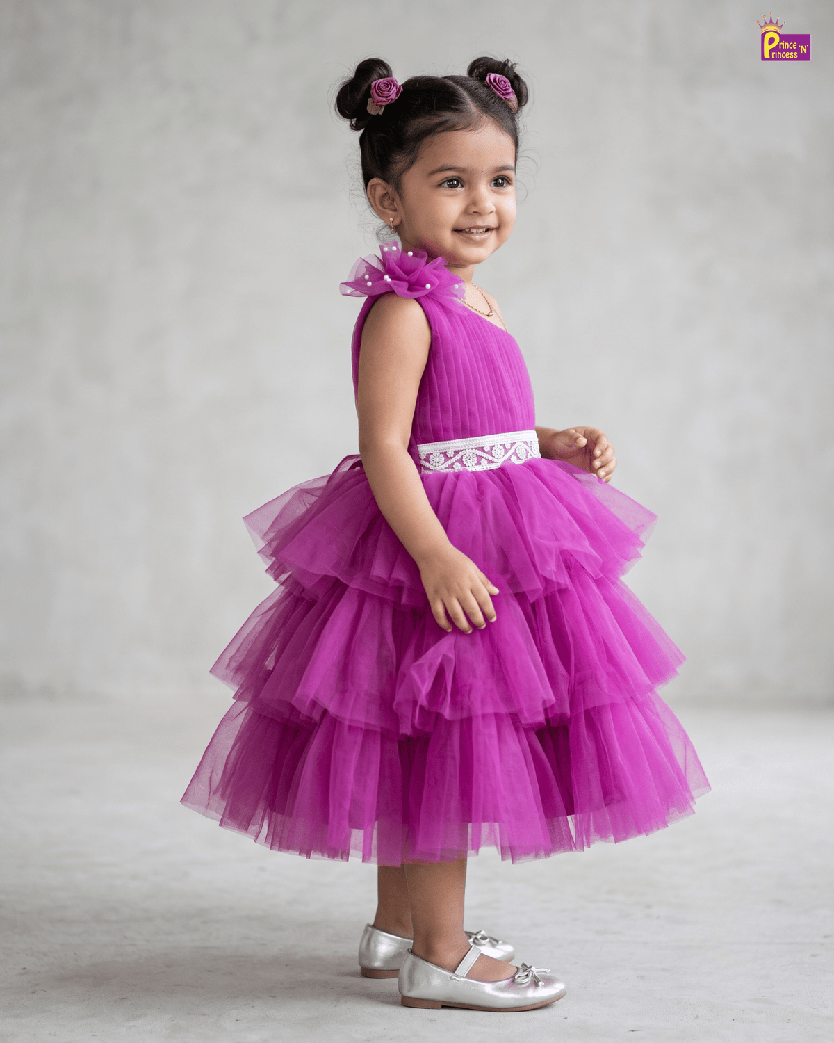 A young girl with dark hair in two buns wearing a bright magenta tiered tulle dress with a white embroidered waist sash, standing against a neutral gray studio background.