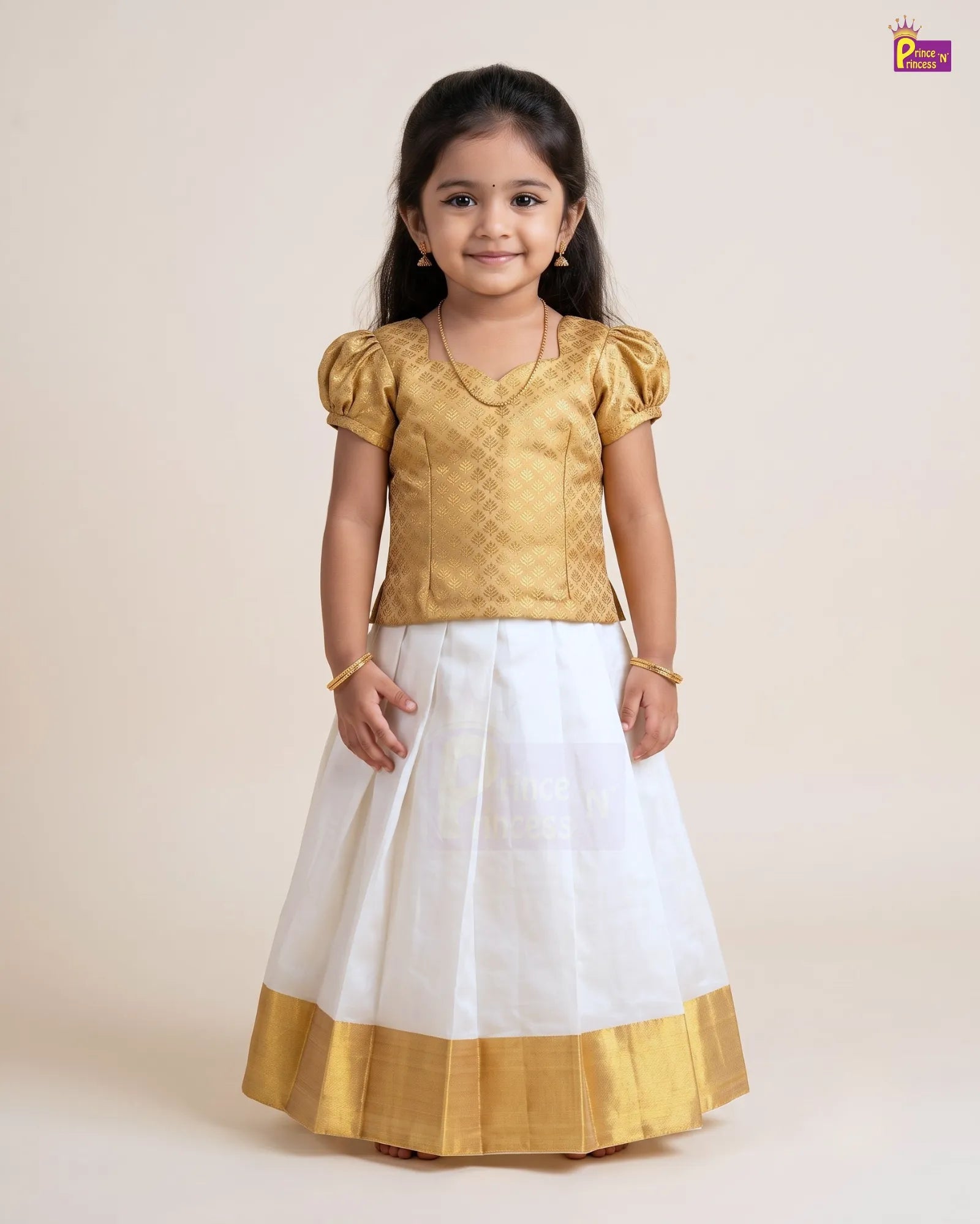A young girl modeling a traditional Indian outfit consisting of a gold metallic embroidered top with puff sleeves and a white skirt with a gold border, set against a plain beige background.