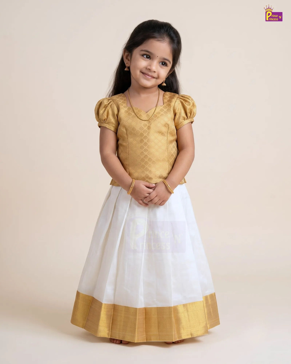 A young girl modeling a traditional Indian dress with a gold textured blouse and a white skirt featuring a gold border, set against a plain beige studio background.