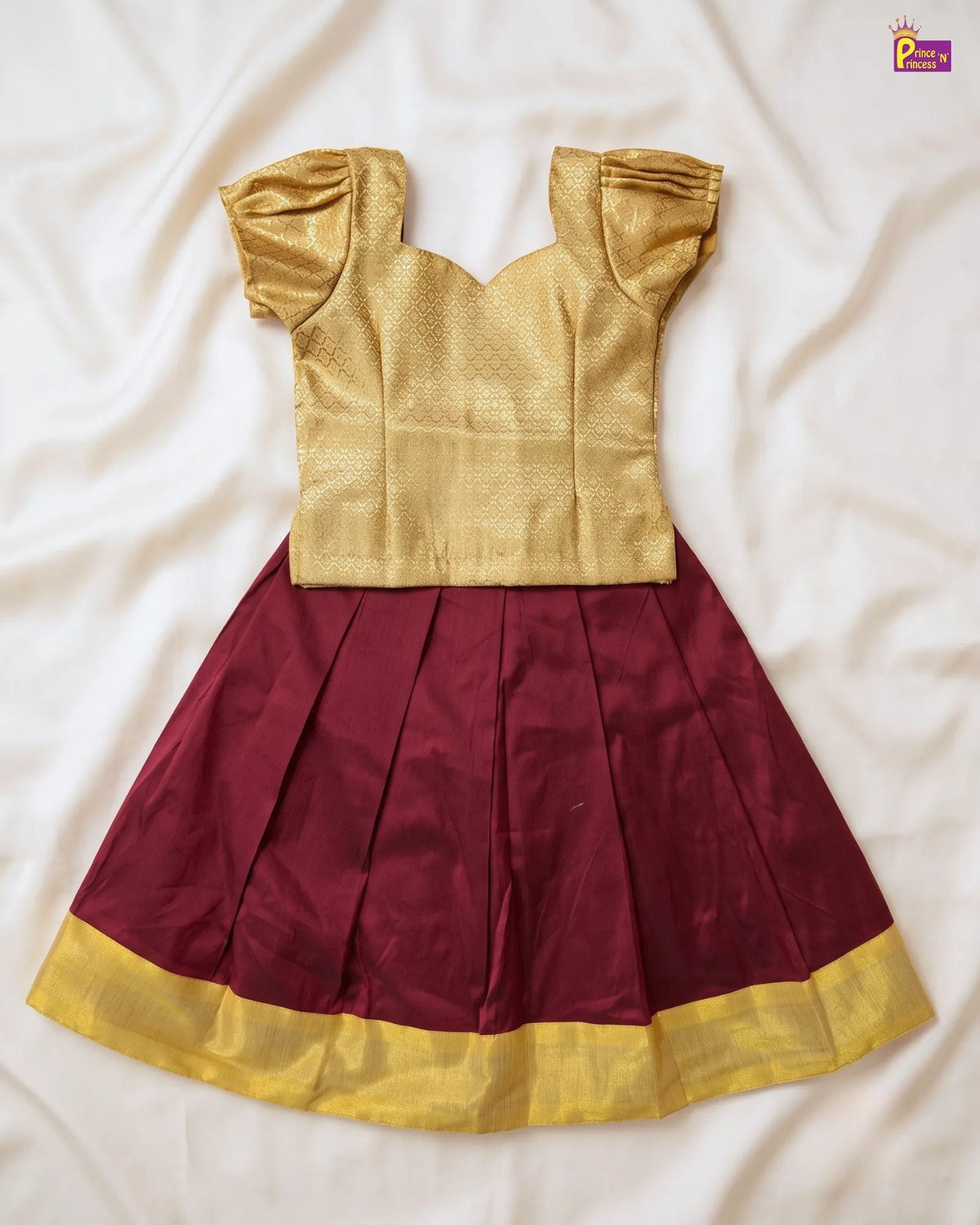 A traditional Indian girl's dress featuring a gold metallic fabric top with puffed sleeves and a pleated maroon skirt with a gold border, laid flat on a white satin sheet.