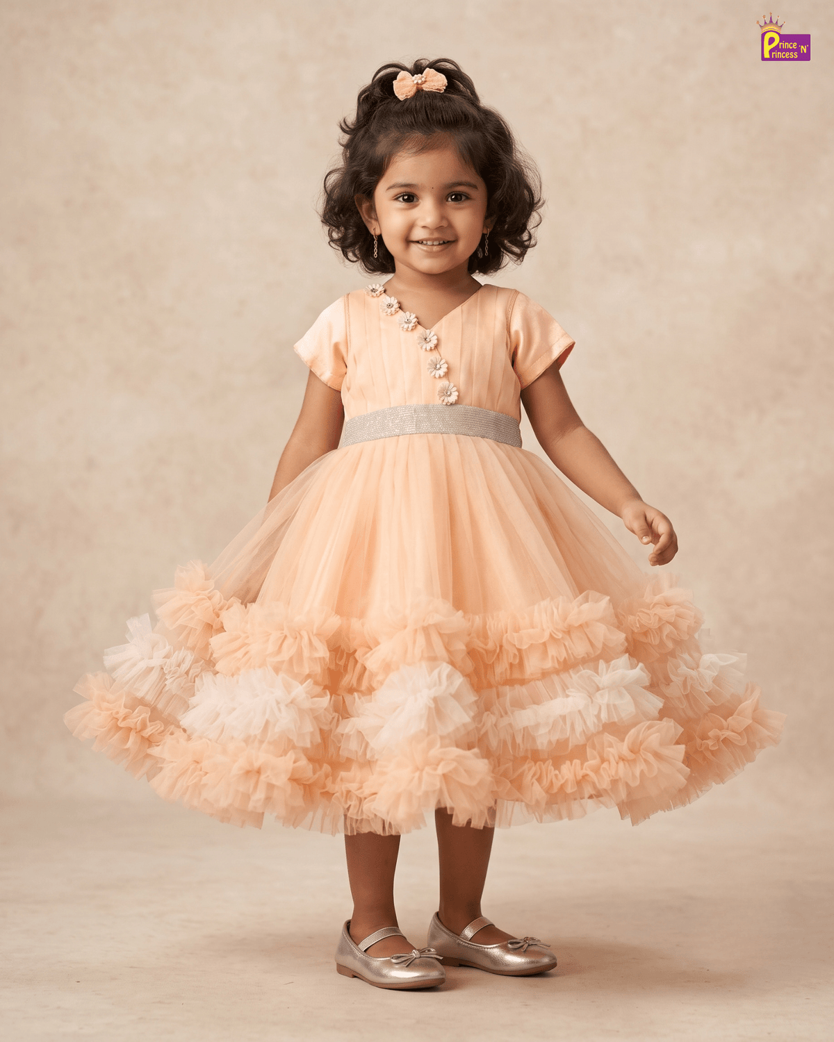 A toddler girl wearing a peach and beige tiered tulle party dress with a lavender waistband and silver shoes, standing against a neutral beige studio backdrop.