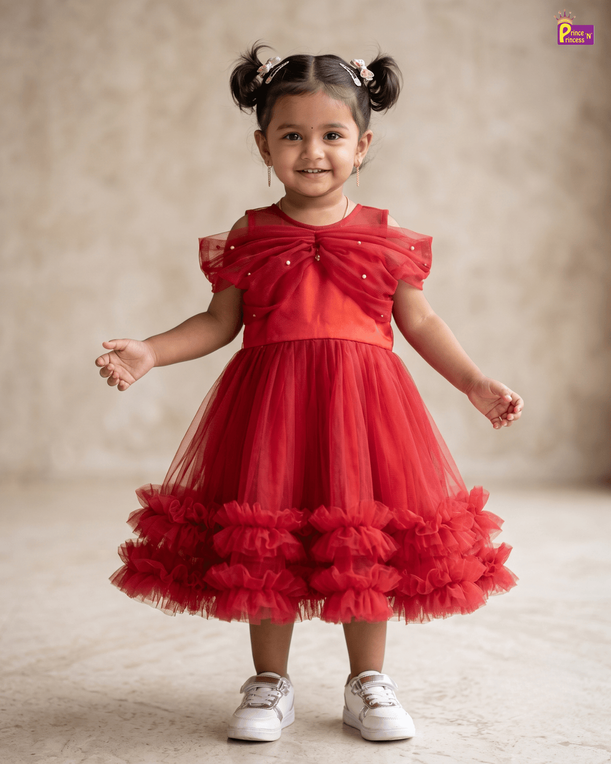 A toddler girl with her hair in two buns wearing a bright red party dress with a ruffled tulle skirt and white sneakers, standing against a neutral beige studio backdrop.