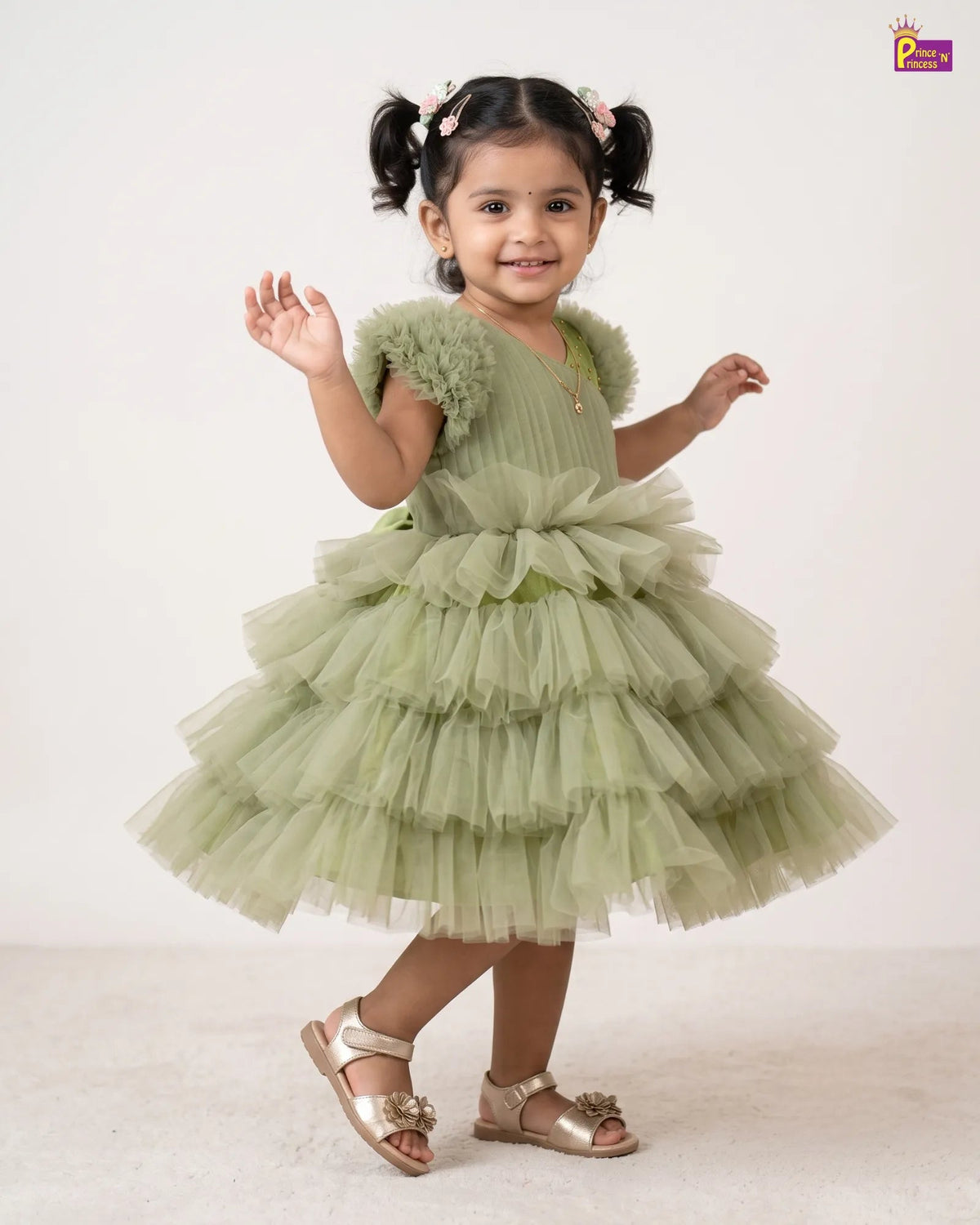 A young girl with dark hair in pigtails models a light sage green tiered tulle party dress with ruffled shoulder details, paired with metallic gold sandals, against a plain off-white studio background.