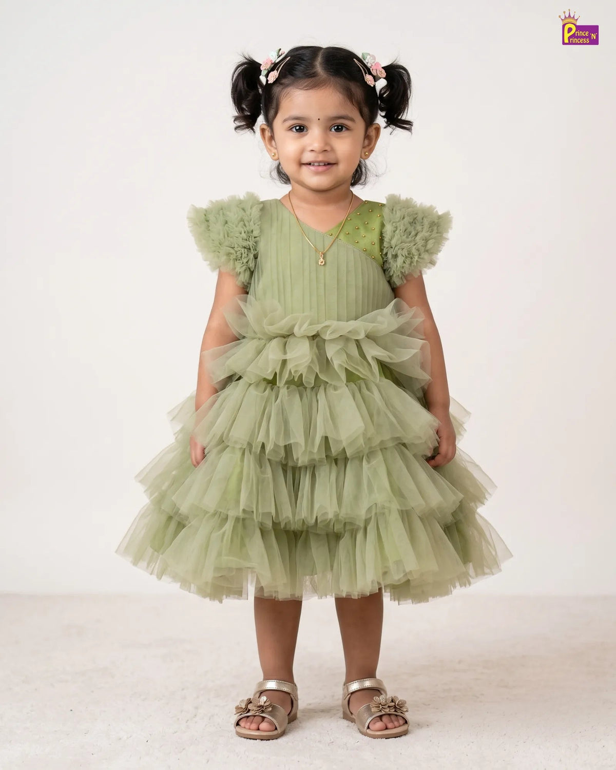 A young girl with dark hair in pigtails stands wearing a light sage green tiered tulle dress with ruffled shoulder details and metallic sandals, photographed against a plain off-white studio background.