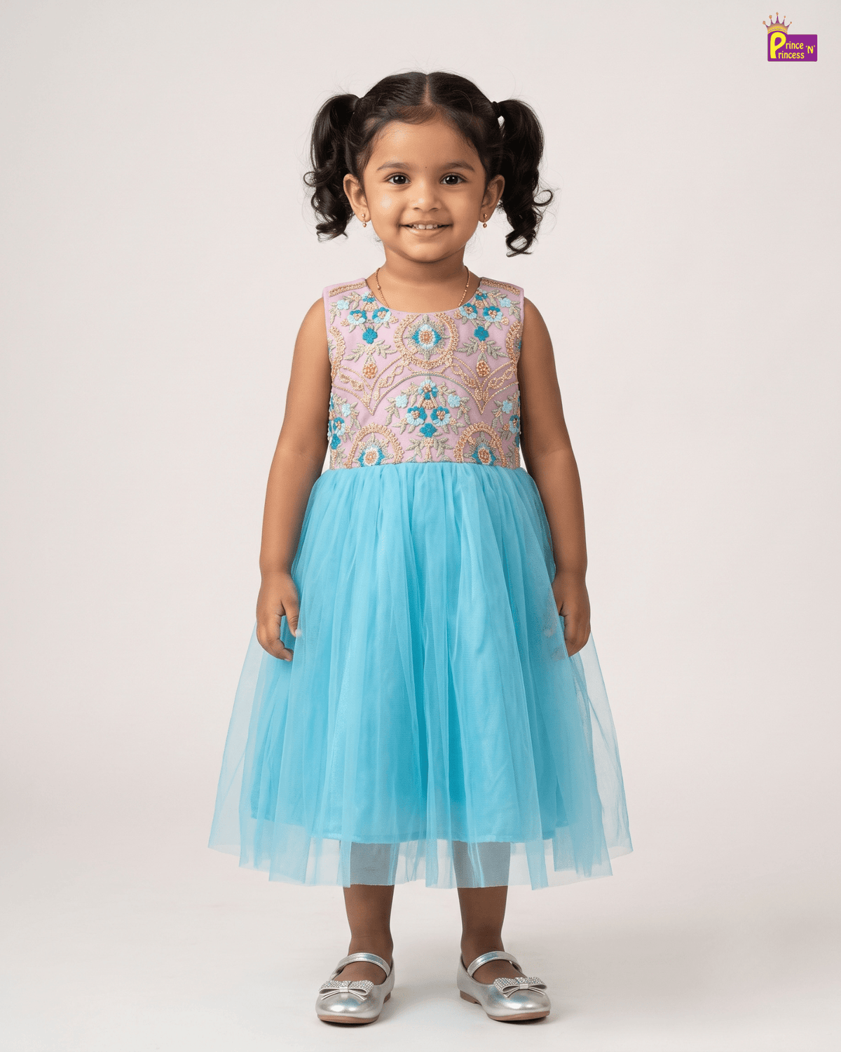 A young girl with dark curly hair in pigtails stands against a light beige studio background wearing a sleeveless tulle dress with a multicolored embroidered bodice and a light blue tulle skirt, paired with silver shoes.