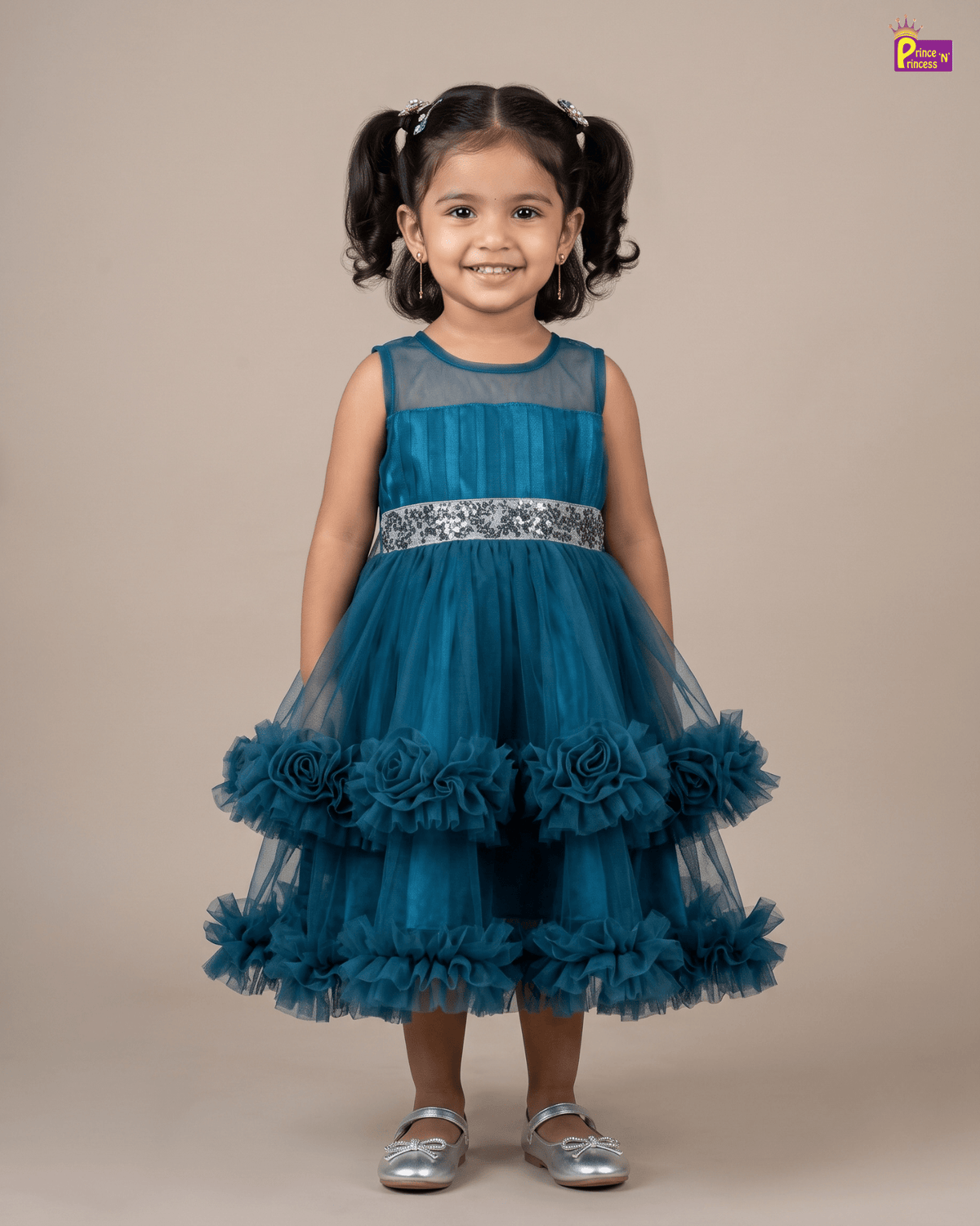 A young girl with dark hair in pigtails stands against a solid beige studio background wearing a sleeveless teal blue tulle dress with a silver patterned waistband and layered ruffle details on the skirt, paired with metallic silver shoes.