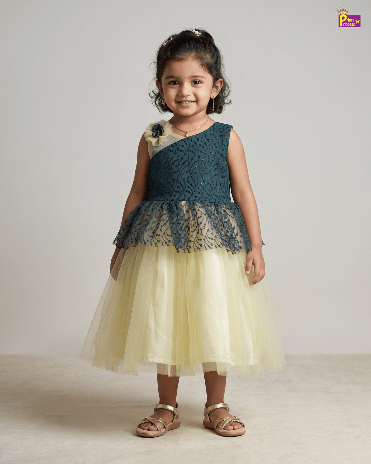 A young girl stands against a plain light gray studio background wearing a sleeveless party dress with a dark teal lace bodice, a pale yellow tulle skirt, and a small floral embellishment on one shoulder. She is also wearing metallic sandals.