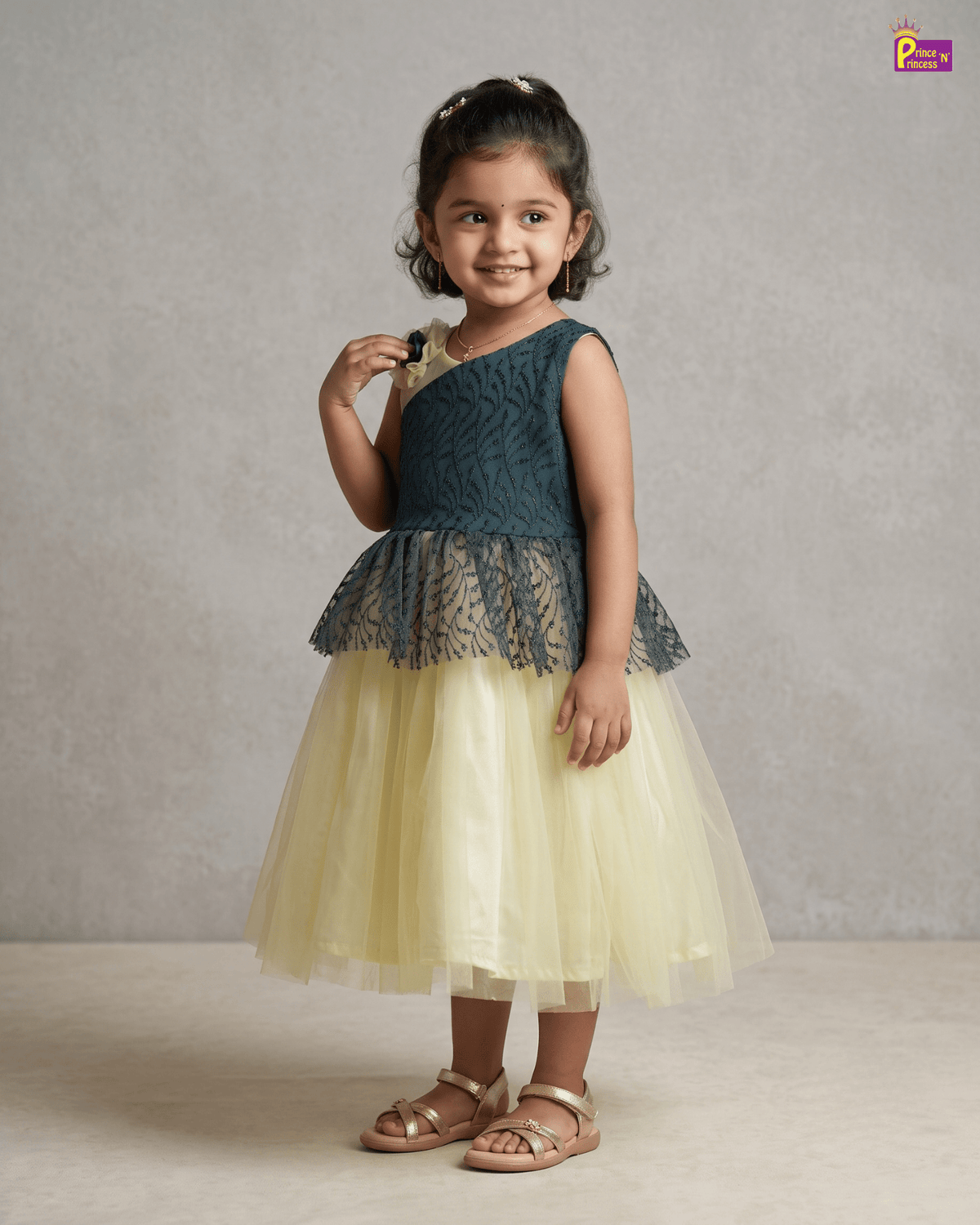 A young girl modeling a sleeveless two-tone dress with a dark teal lace bodice and a pale yellow tulle skirt, paired with gold sandals, set against a neutral textured studio backdrop.