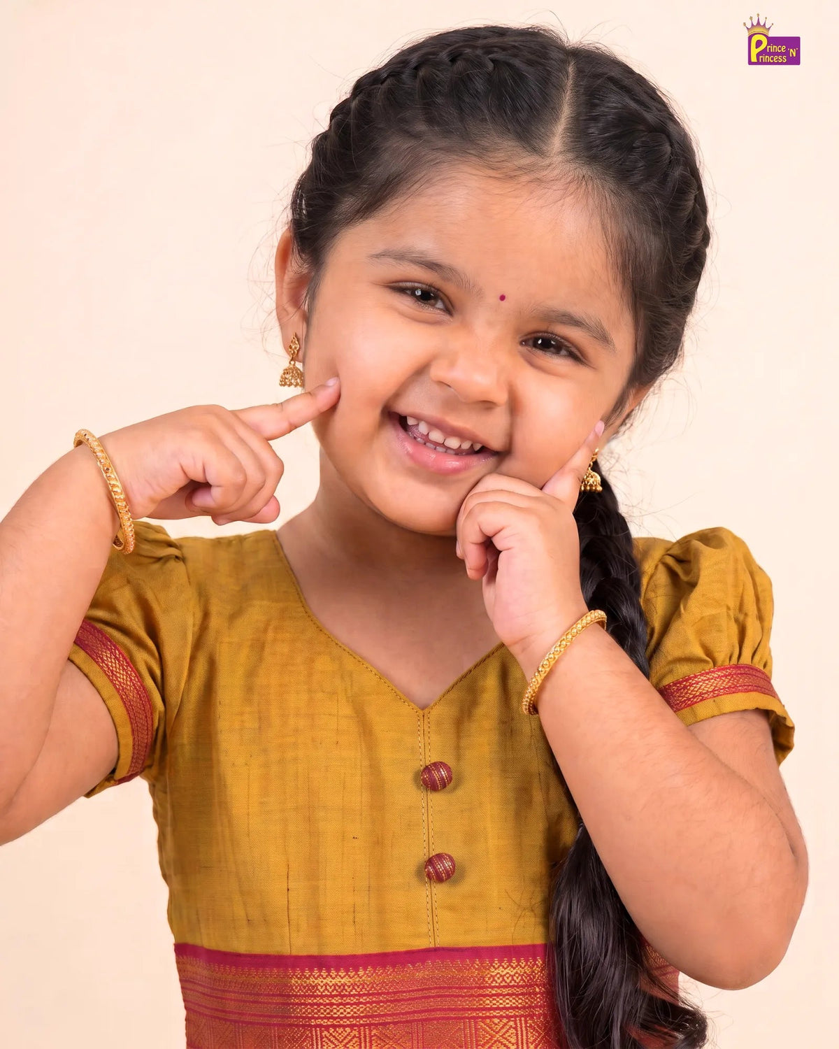 A young girl with dark braided hair smiles while pointing at her cheeks, wearing a mustard yellow traditional outfit with red borders and gold jewelry against a plain beige background.