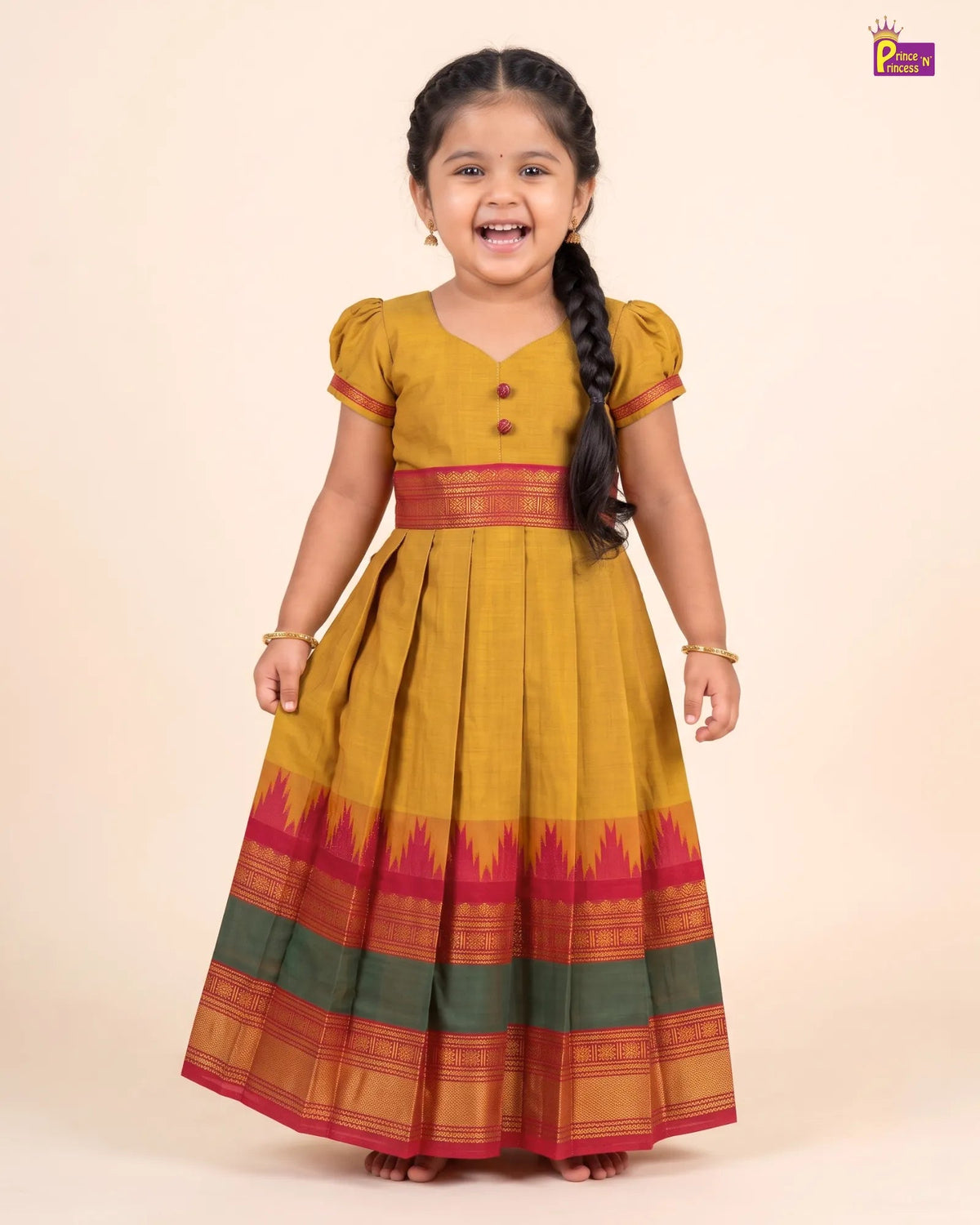 A young girl with a long black braid is wearing a traditional Indian dress, standing against a solid beige background. The dress is mustard yellow with a red waistband and a wide border at the bottom featuring red, green, and gold patterns.