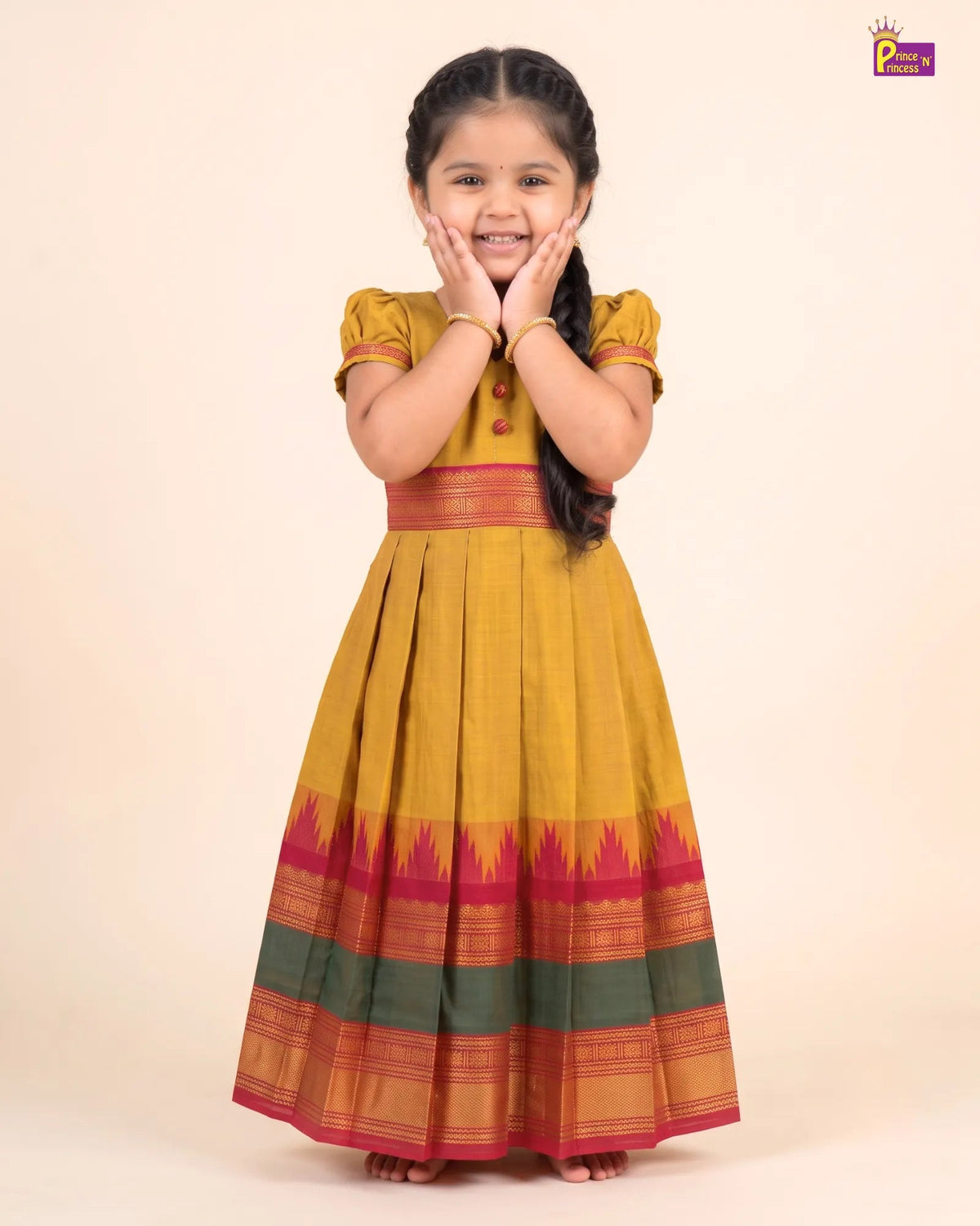 A young girl with braided hair stands barefoot against a plain beige studio background, wearing a traditional long dress with a mustard yellow body, a red waistband, and a flared skirt with red, orange, and green patterned bands. She is smiling with her hands near her face.