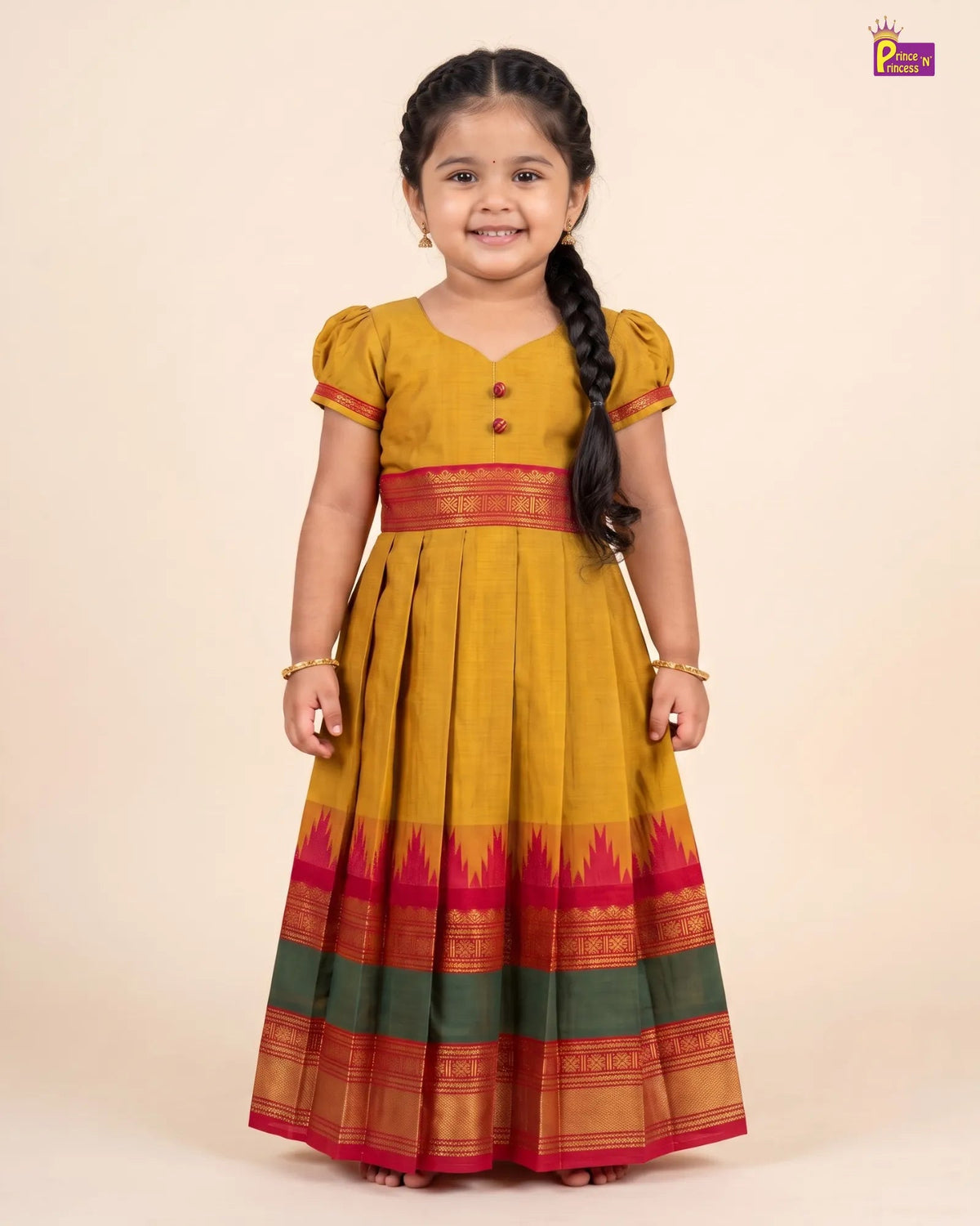 A young girl with a long black braid stands wearing a traditional Indian mustard yellow cotton dress with red and green borders, set against a solid beige background.