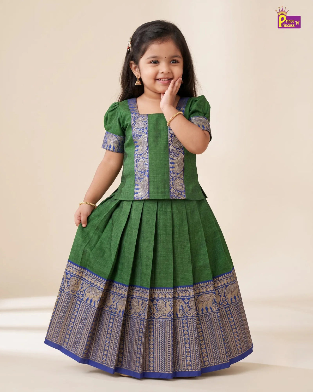 A young girl modeling a green traditional Indian dress with blue and gold patterned borders and embroidery, standing against a plain beige studio background.