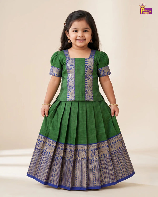 A young girl modeling a green and blue traditional Indian dress with intricate gold and blue patterns, set against a plain beige background.