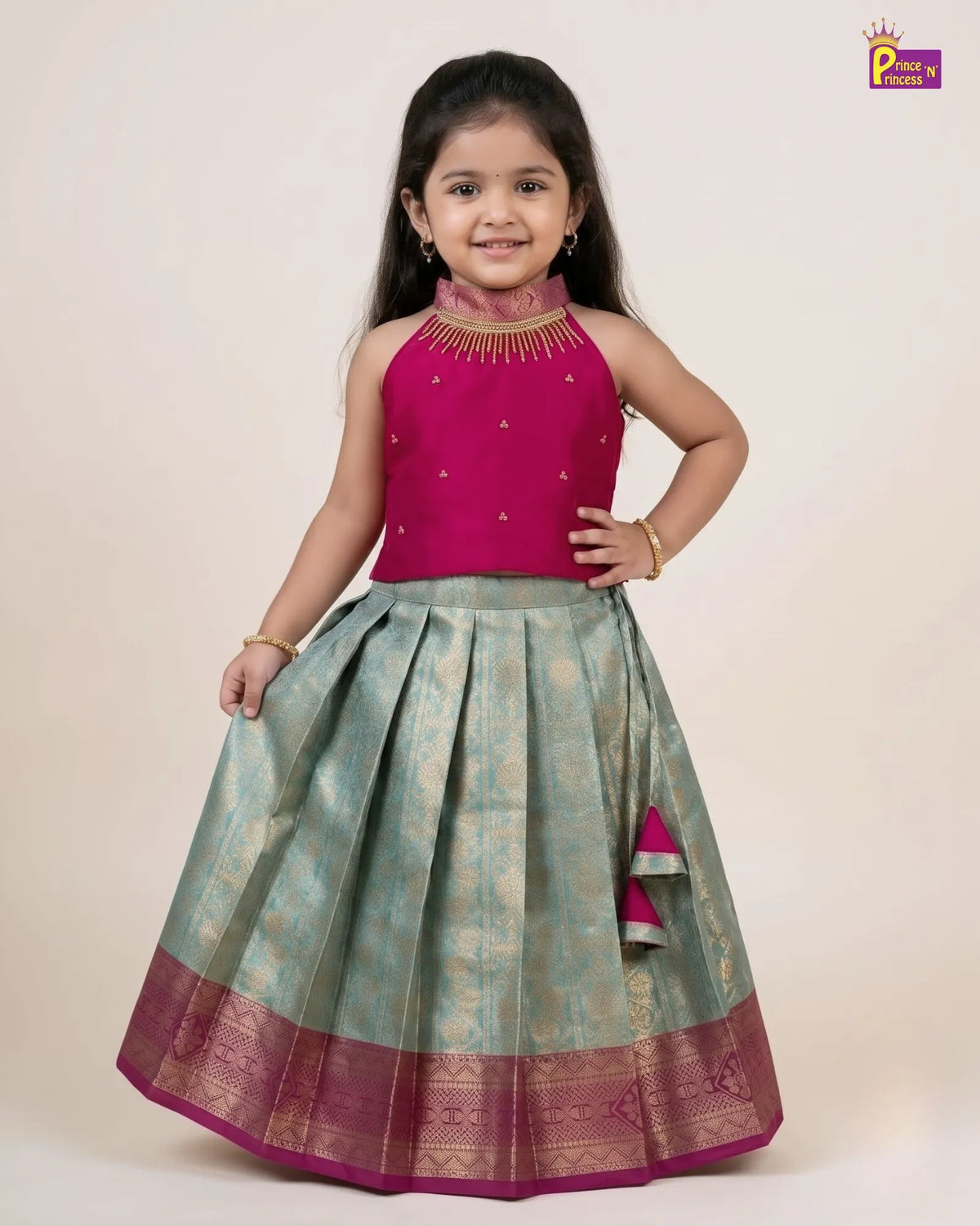 A young girl modeling a traditional Indian ethnic wear set consisting of a magenta pink sleeveless top with gold embroidery and a matching pleated light green skirt with a magenta border, on a plain beige background.