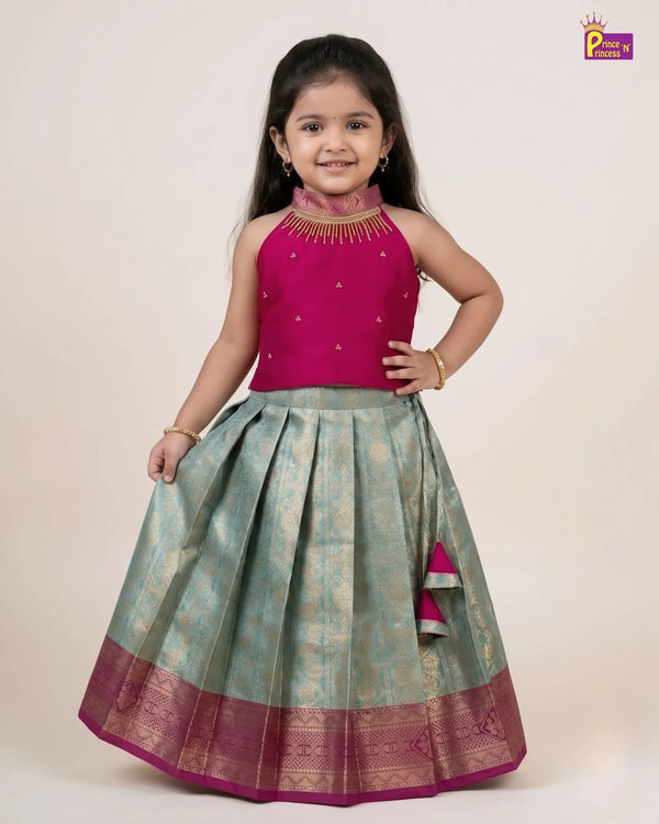 A young girl modeling a traditional Indian ethnic wear set consisting of a magenta pink sleeveless top with gold embroidery and a matching pleated light green skirt with a magenta border, on a plain beige background.