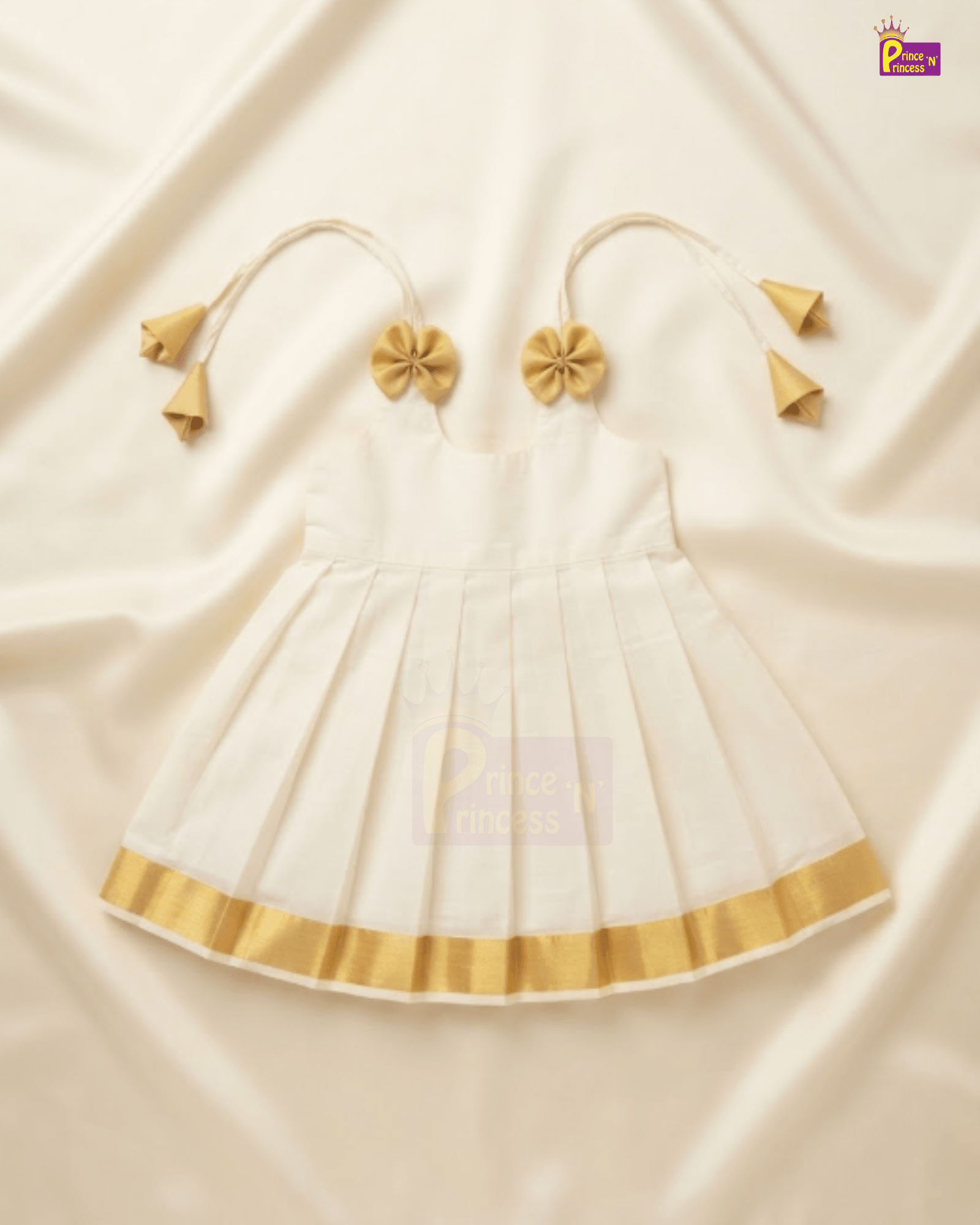 A white baby girl's dress with gold trim and matching gold tie-shoulder accessories, laid flat on a cream-colored satin fabric background.