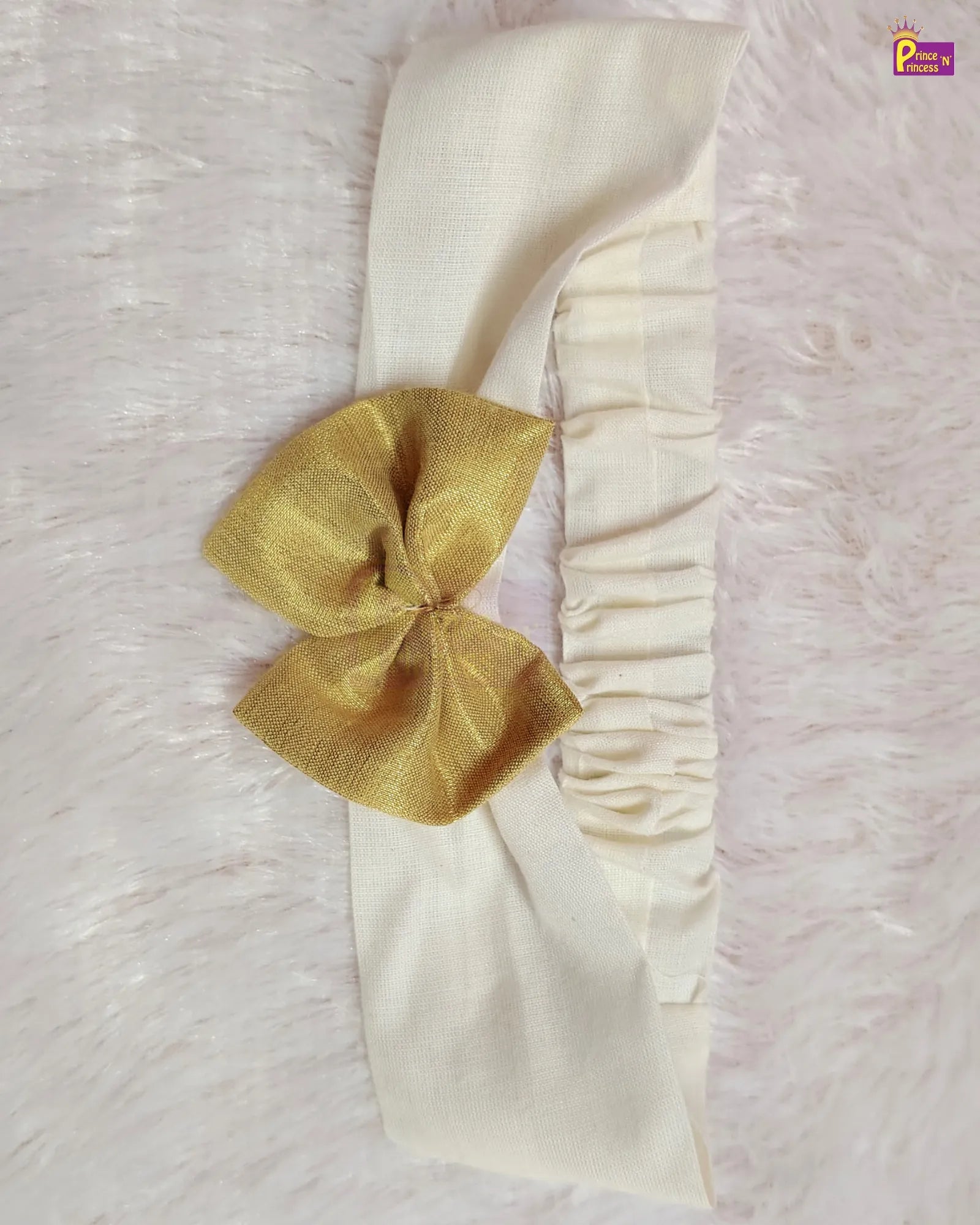 An off-white fabric headband with a gathered elastic band and a large gold satin bow, displayed on a white faux fur background.