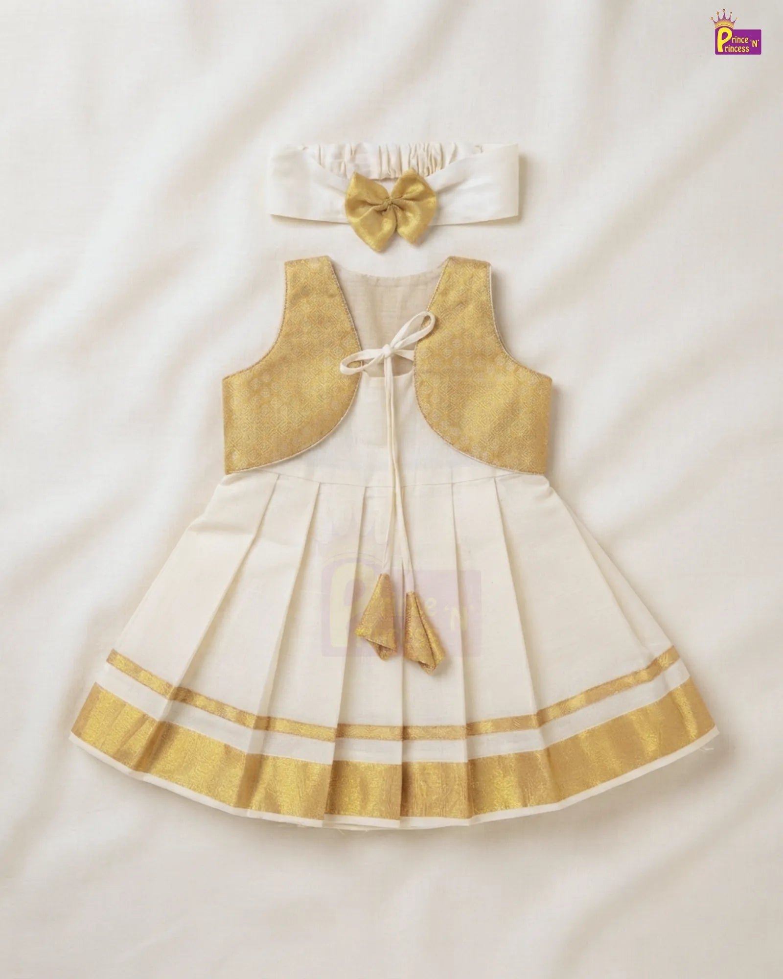 A toddler girl's traditional Indian outfit set featuring a cream-colored dress with gold borders, a matching gold vest, and a white headband with a gold bow, laid flat on a white fabric background.