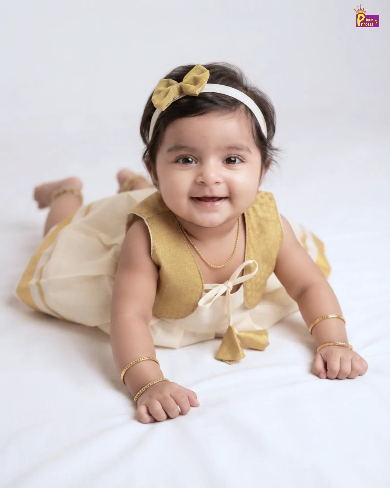 A baby girl lying on her stomach wearing a cream and mustard yellow dress with a matching yellow bow headband and gold bangles, set against a plain white studio background.