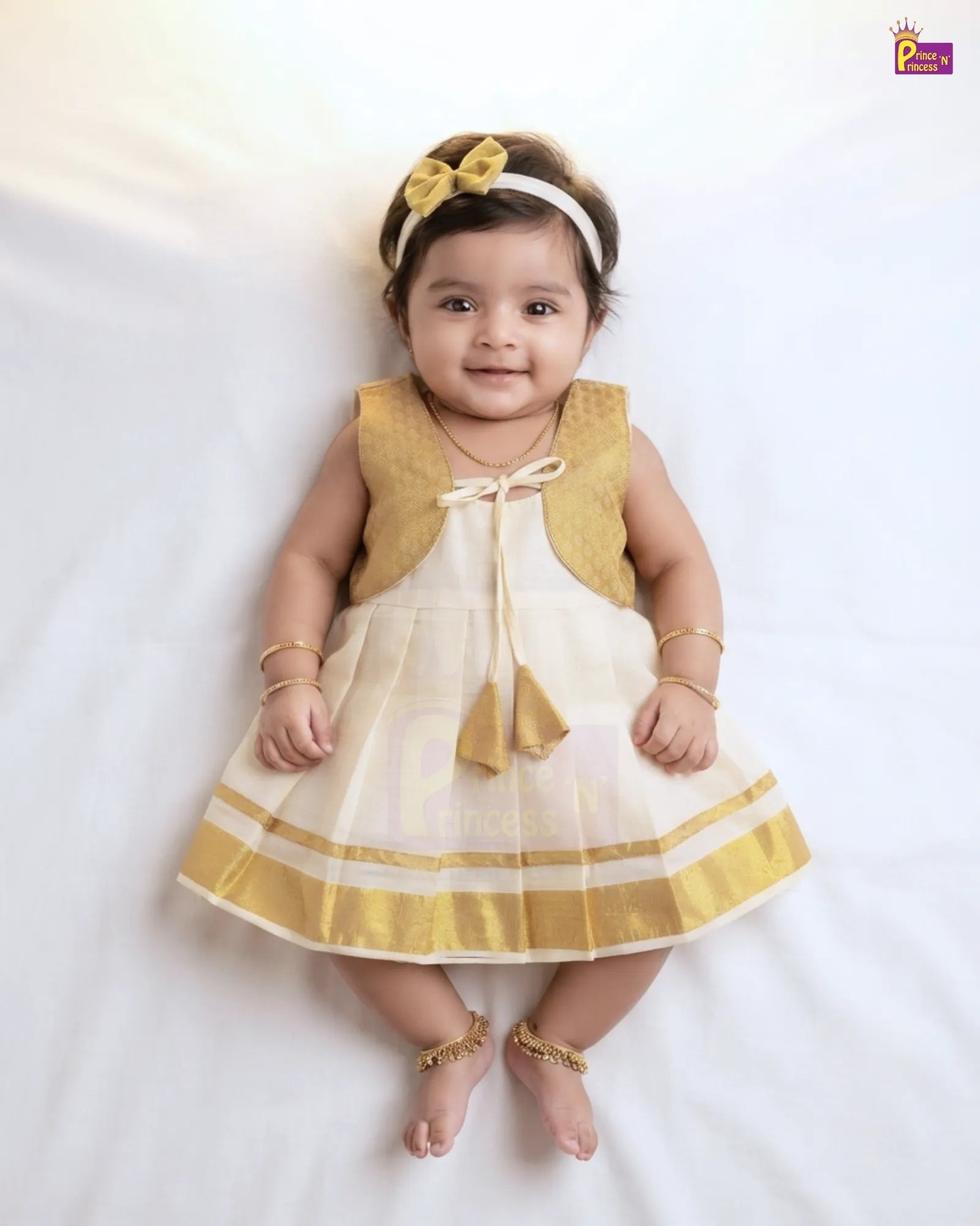 A baby girl wearing a traditional Indian outfit consisting of a white and gold dress with a gold vest, a gold bow headband, and gold jewelry, lying on a white fabric background.