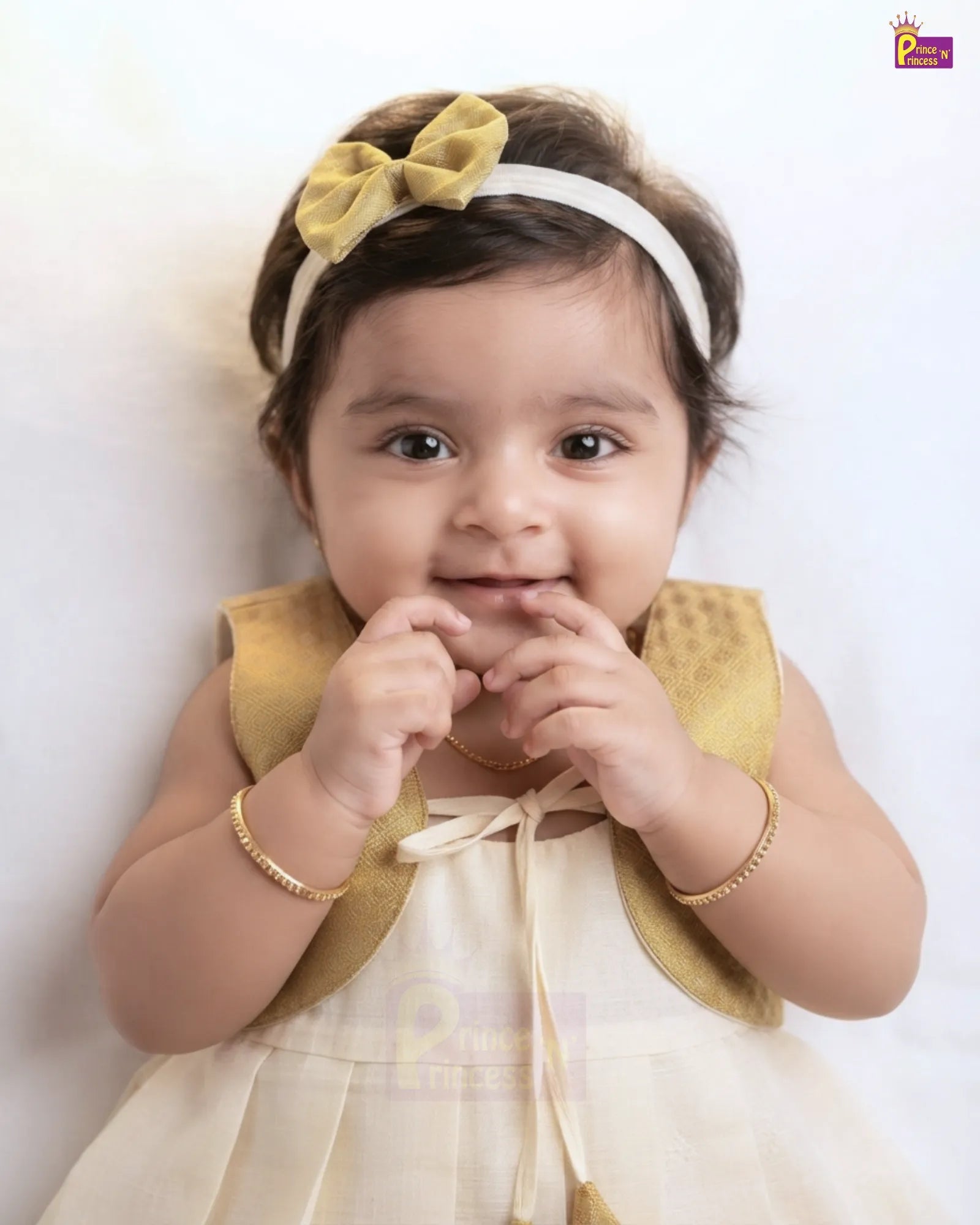 A baby girl wearing a cream-colored dress with a gold mesh collar, a matching gold bow headband, and gold bracelets, lying down against a white background.