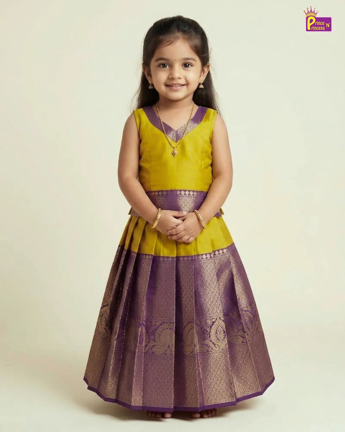 A young girl modeling a traditional Indian dress with a yellow upper bodice and a purple metallic skirt, standing against a plain light beige background.