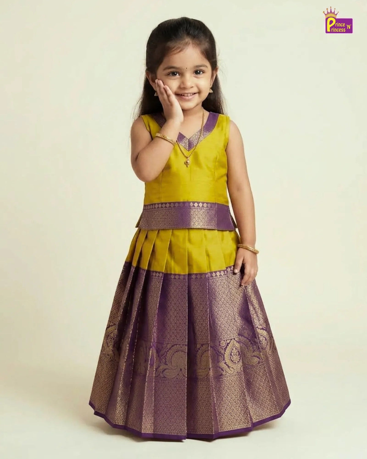 A young girl modeling a traditional Indian ethnic outfit consisting of a yellow sleeveless kurta with a purple border and a matching pleated purple skirt with gold zari embroidery, set against a plain light beige background.
