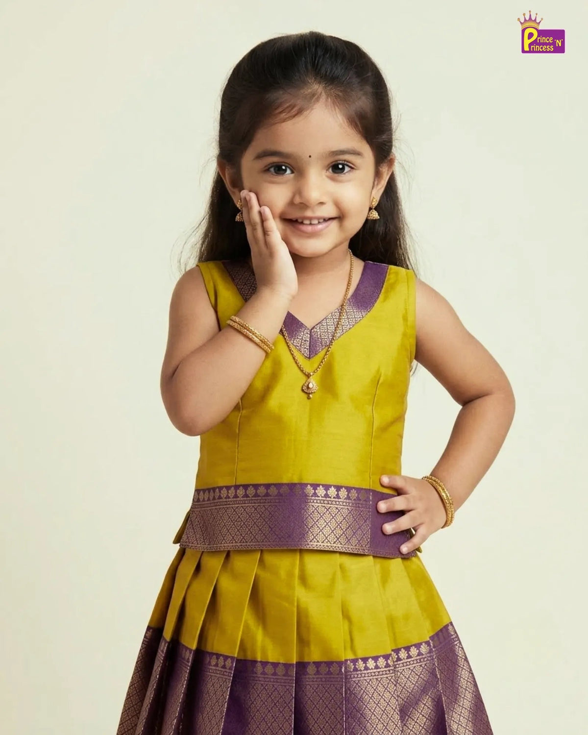 A young girl modeling a traditional Indian dress with a mustard yellow top and a pleated purple skirt with gold zari borders, set against a plain light beige background.
