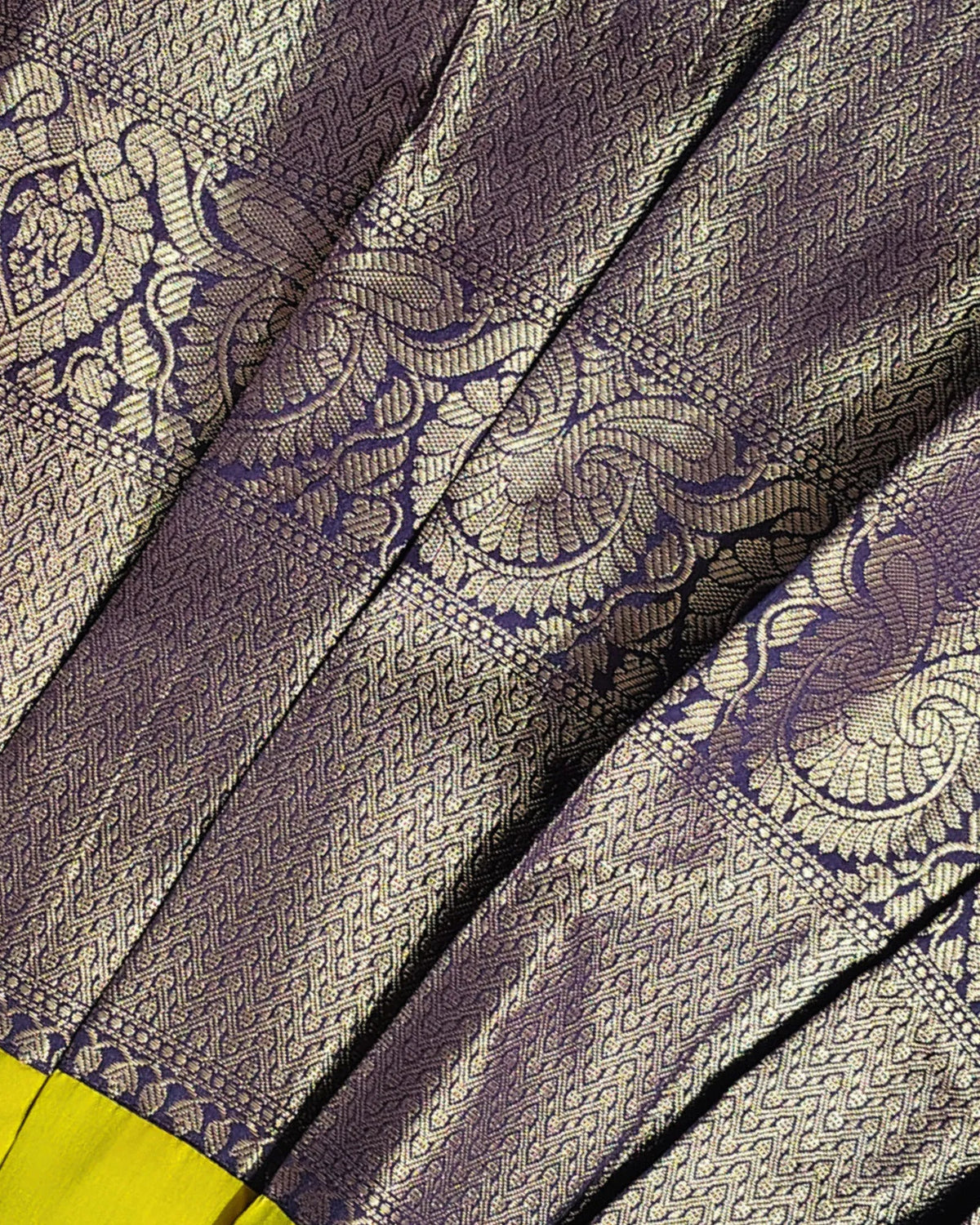 Close-up of a woven fabric with a metallic lavender-purple base and intricate gold zari floral and paisley patterns, showing the texture and sheen of the material.