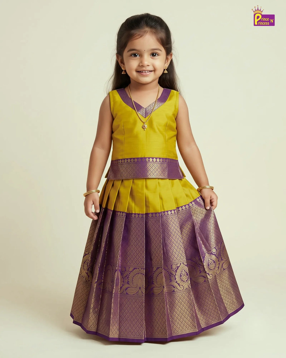 A young girl modeling a traditional Indian ethnic dress with a yellow sleeveless top and a pleated purple skirt with gold zari detailing, set against a plain beige background.