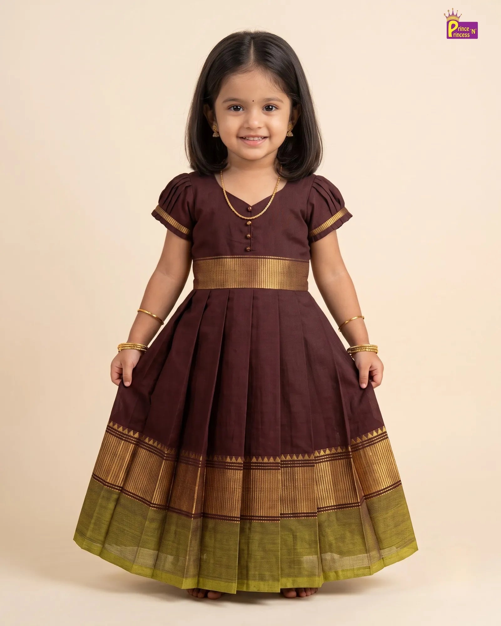 A young girl modeling a dark brown A-line dress with gold and olive green zari borders, standing against a solid beige background.