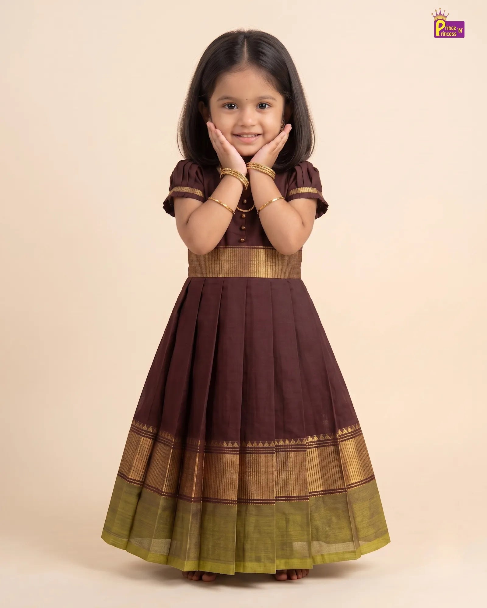 A young girl modeling a traditional Indian salwar kameez dress. The dress features a dark brown bodice with gold zari embroidery, a wide gold waistband, and a pleated skirt with a green and gold border.
