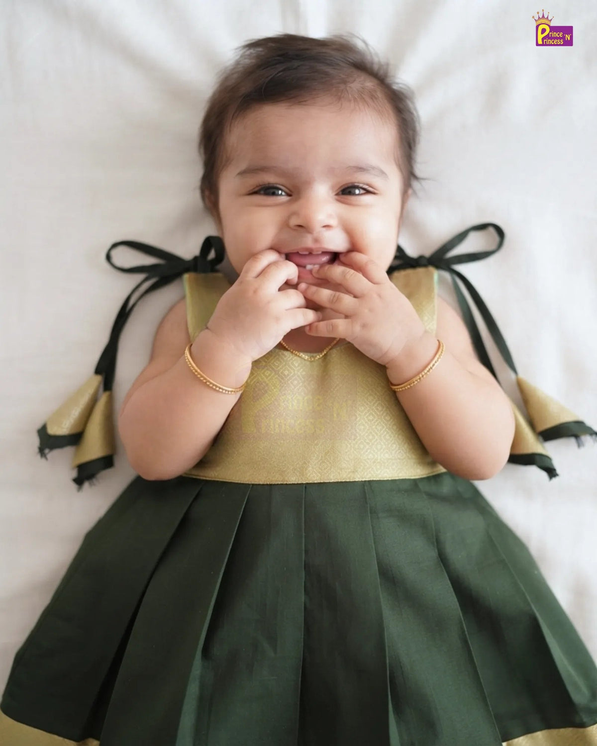 A baby girl lying on a white sheet, wearing a sleeveless green and gold dress with black ribbon bows on the back. She is smiling and touching her mouth, wearing gold bracelets.