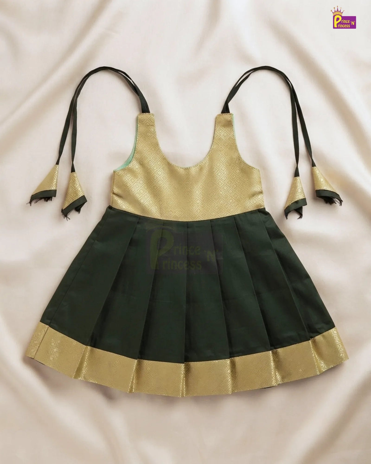 A children's sleeveless dress with a metallic gold upper bodice and a dark green pleated skirt, laid flat on a light beige fabric background. The dress has thin black shoulder ties with gold and black tassels and a gold border along the skirt hem.