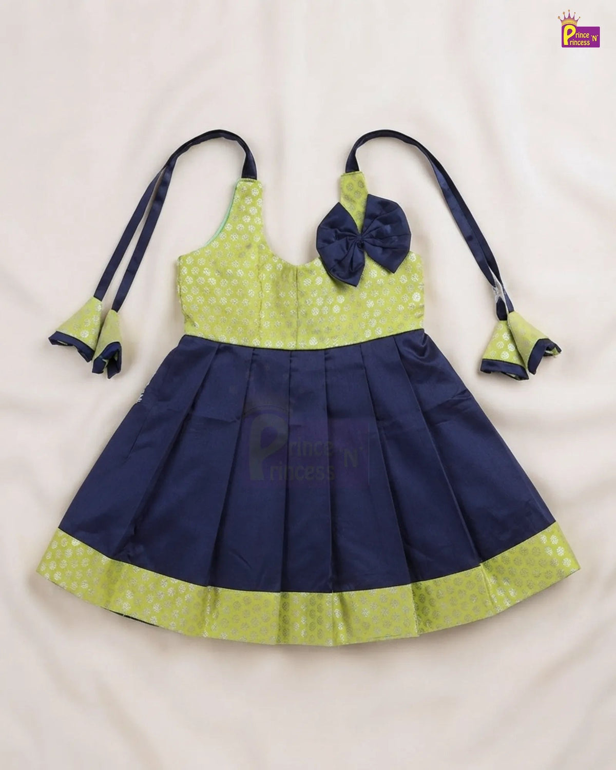 A toddler girl's sleeveless party dress laid flat on a white fabric background. The dress features a lime green and white patterned bodice with a matching bow on the chest, a navy blue pleated skirt, and matching navy blue tie straps with ruffled trim.
