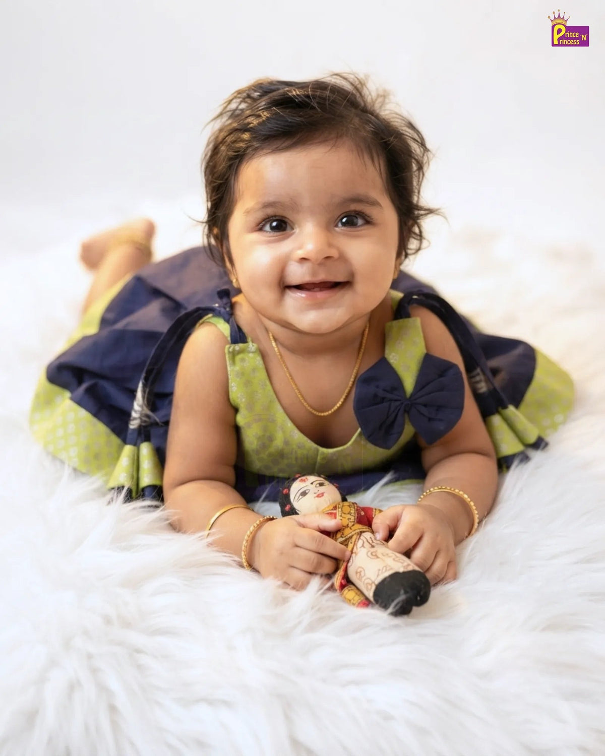 A smiling toddler lying on a white faux fur rug, wearing a green and navy blue dress with a large navy bow on the chest and holding a small doll.