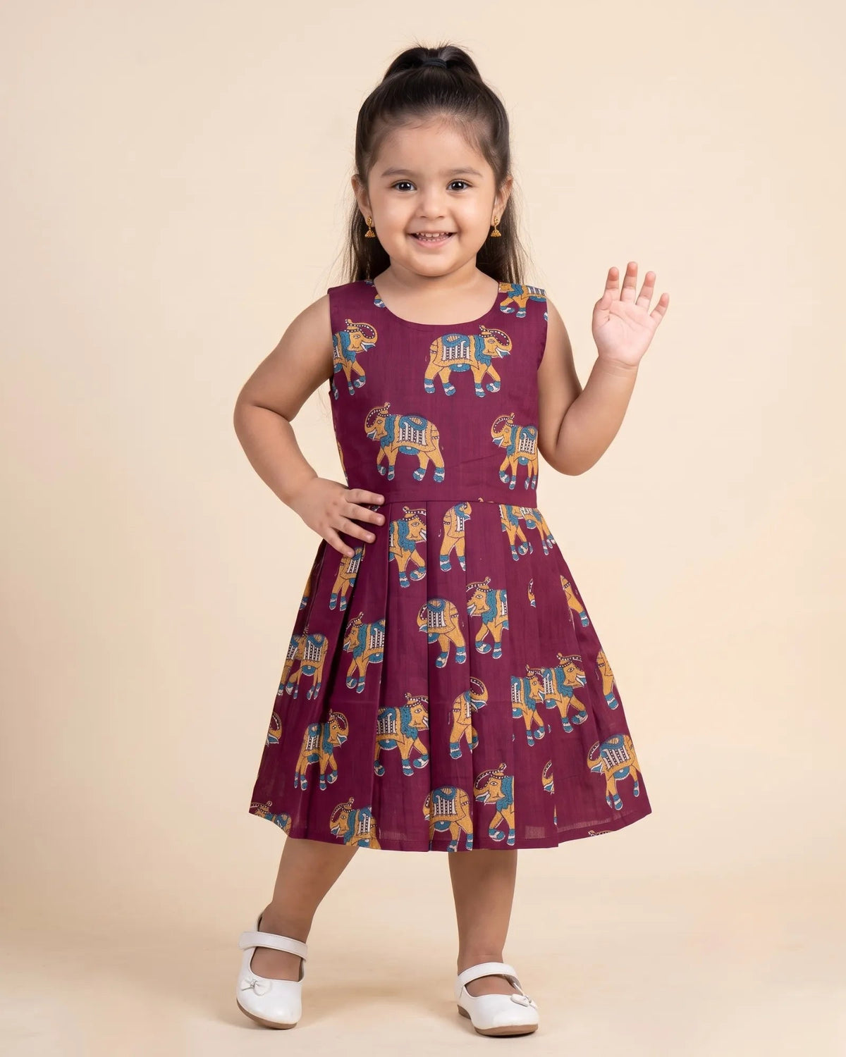 A young girl with her hair in a high ponytail stands smiling and waving, wearing a sleeveless maroon dress with a colorful elephant print and white Mary Jane shoes, set against a solid beige studio background.