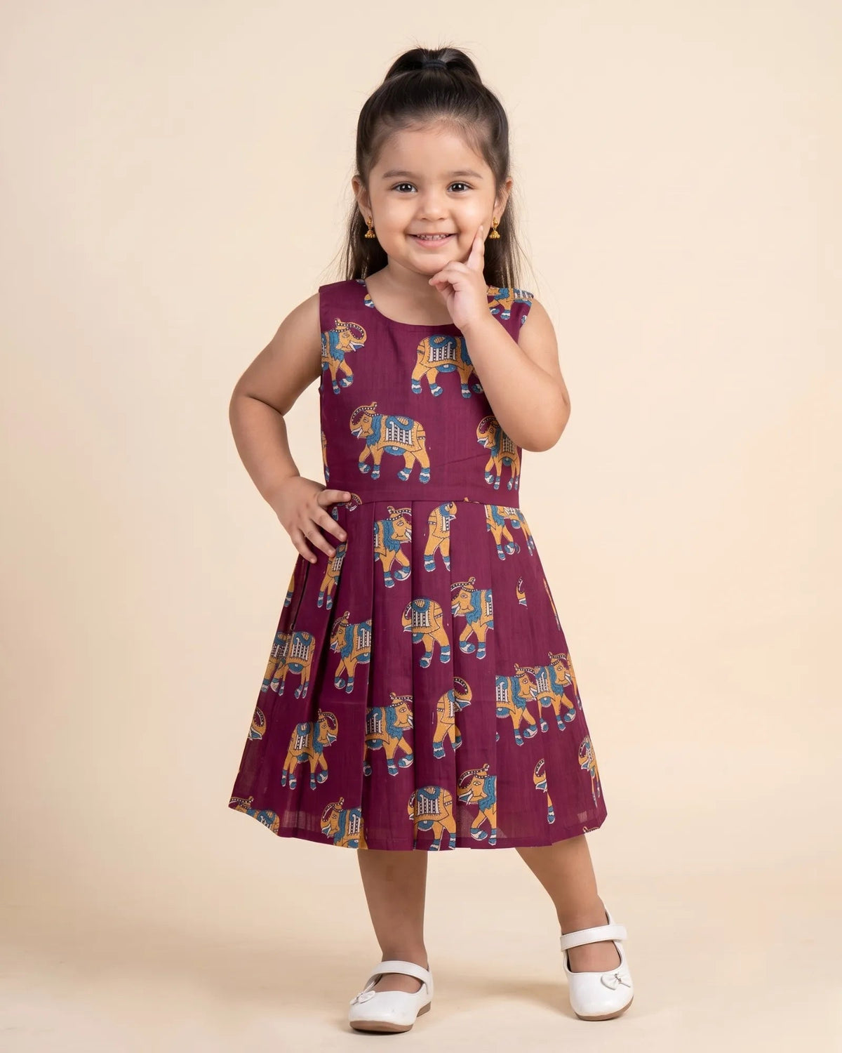 A young girl with her hair in a high ponytail stands in a studio wearing a sleeveless maroon dress with a repeating elephant print in blue, yellow, and white. She is wearing white Mary Jane shoes against a solid light beige background.