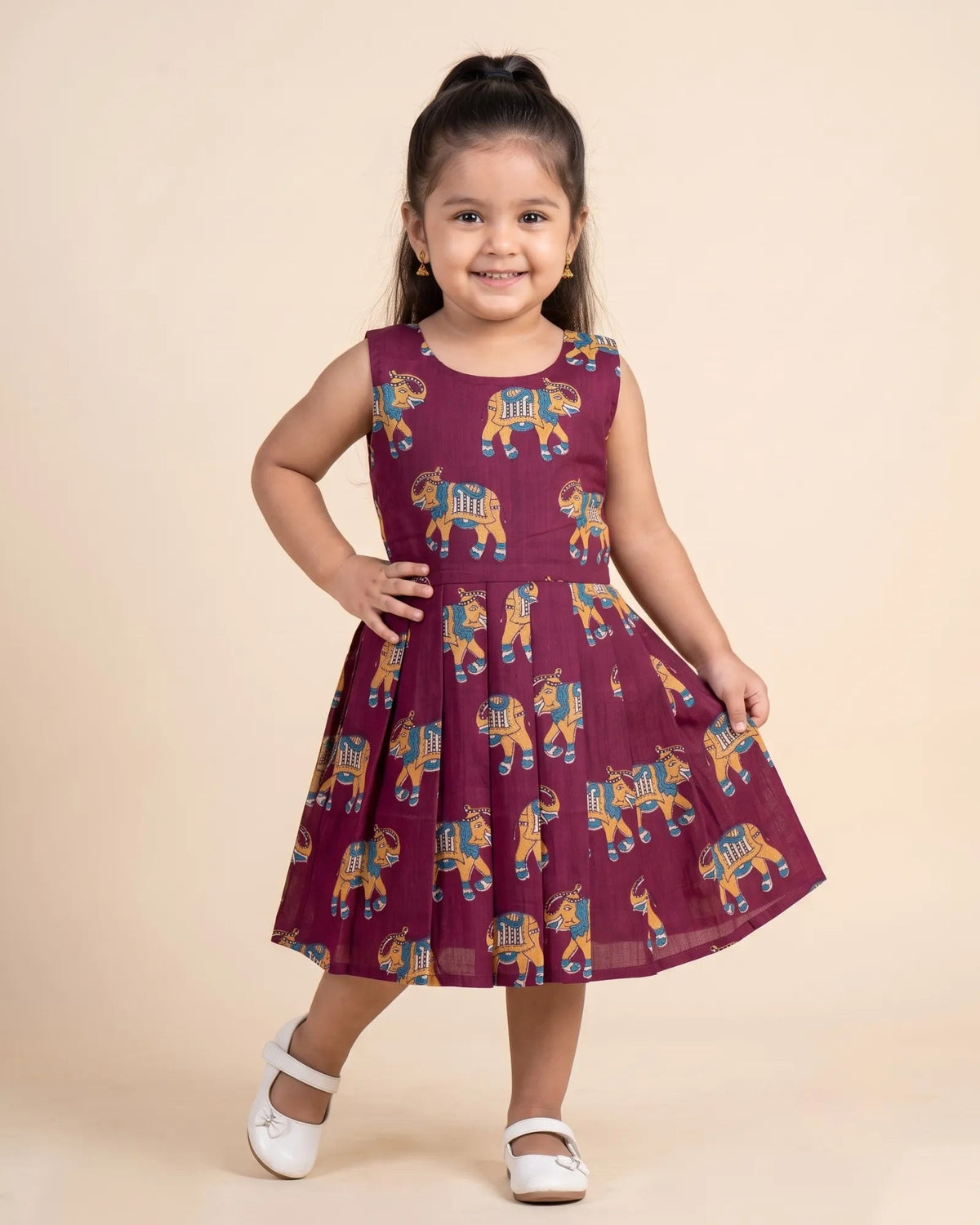 A young girl with her hair in a high ponytail stands smiling in a sleeveless maroon dress with a colorful elephant print, paired with white shoes, against a solid beige studio background.