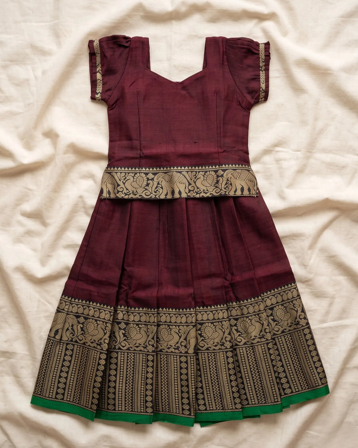 A maroon cotton anarkali dress with gold zari embroidery, featuring a flared skirt and a green hem, laid flat on a cream-colored fabric background.