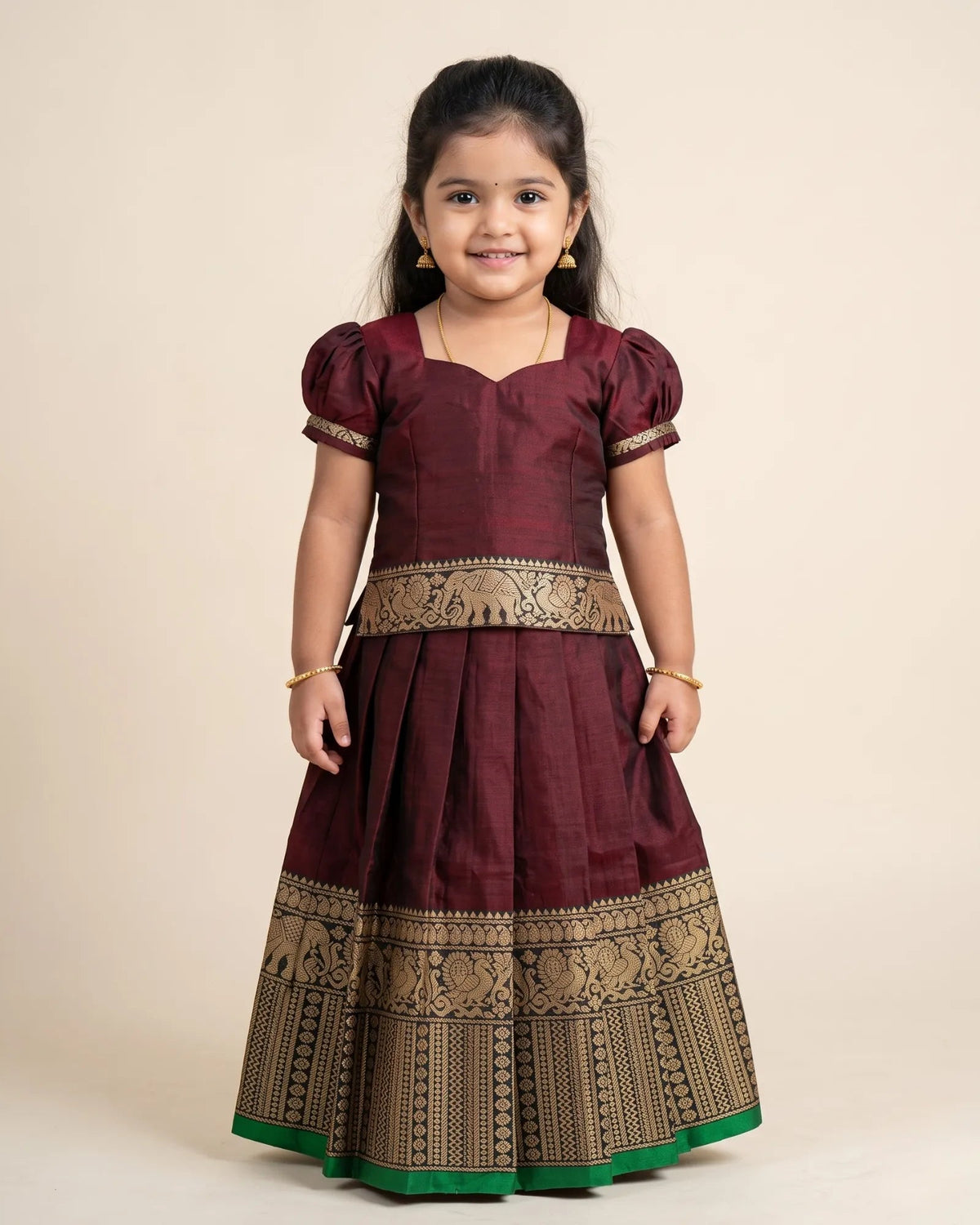 A young girl wearing a traditional Indian maroon and gold Banarasi dress with a green border, standing against a plain beige background.