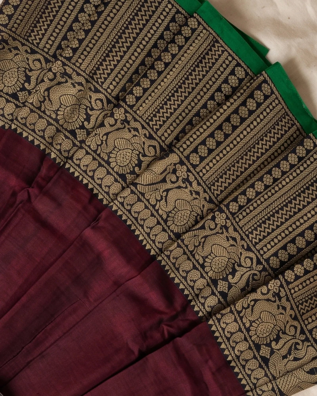 A close-up of a traditional Indian saree fabric, showing a deep maroon body, a wide black and gold patterned border with intricate floral and geometric motifs, and a glimpse of a green border.