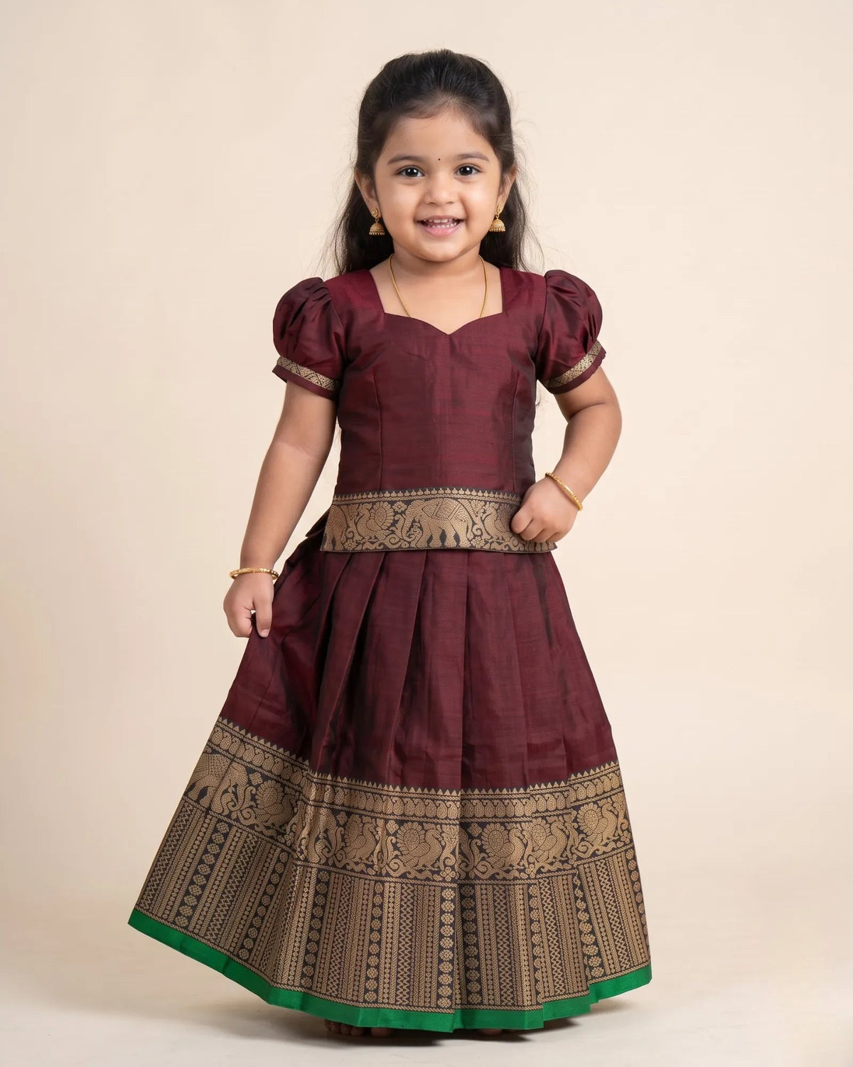 A young girl modeling a traditional Indian dress, featuring a deep maroon body with a beige patterned waistband and a wide border on the skirt, finished with a green hem, set against a plain beige background.