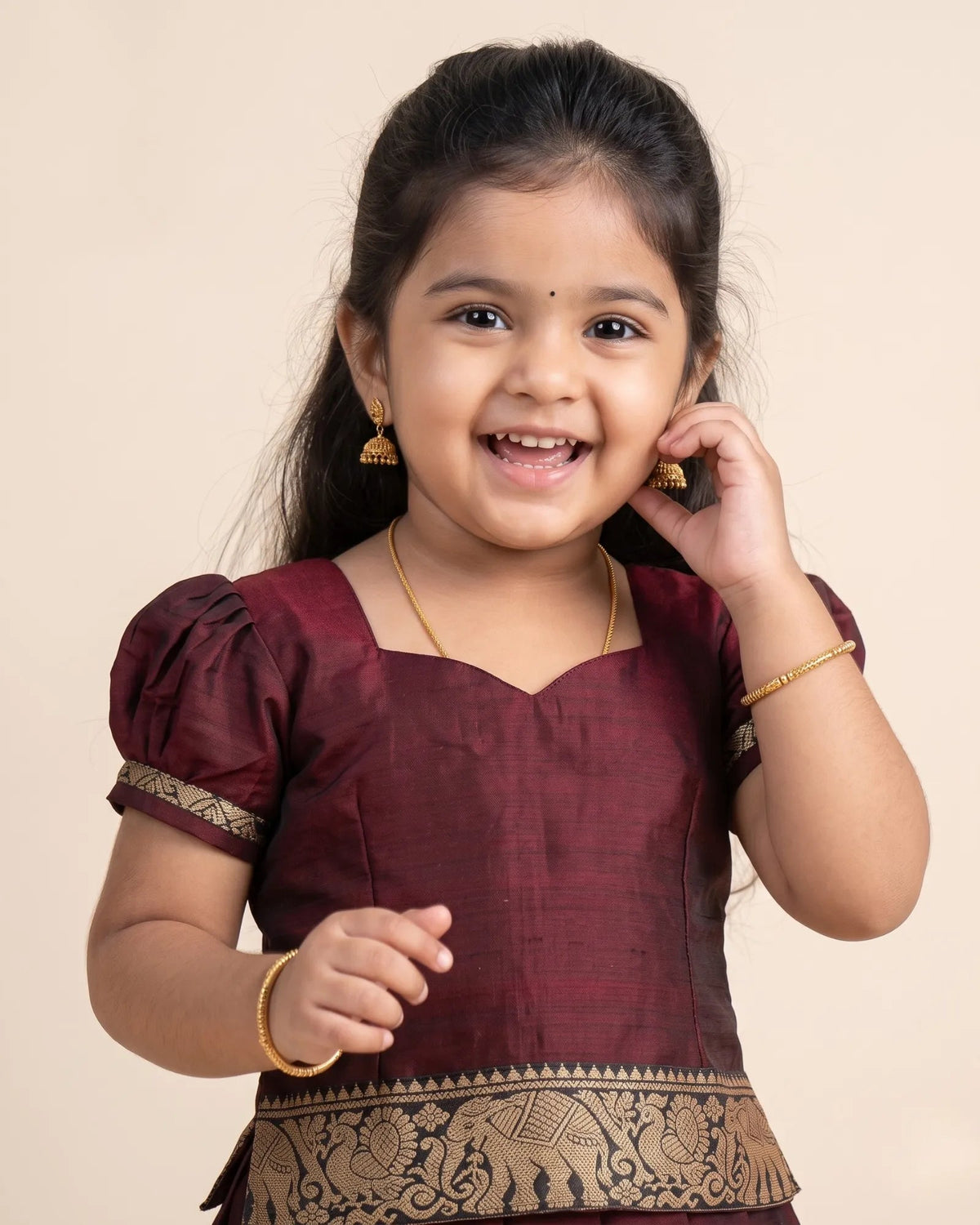A young girl smiling and posing in a traditional maroon and gold embroidered Indian outfit against a solid beige background.