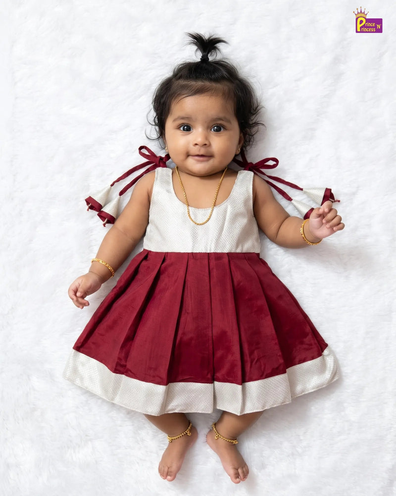 A baby girl lying on a white textured blanket, wearing a sleeveless red and white dress with pleated skirt, white shoulder panels, and red ribbon details tied at the back of the neck.